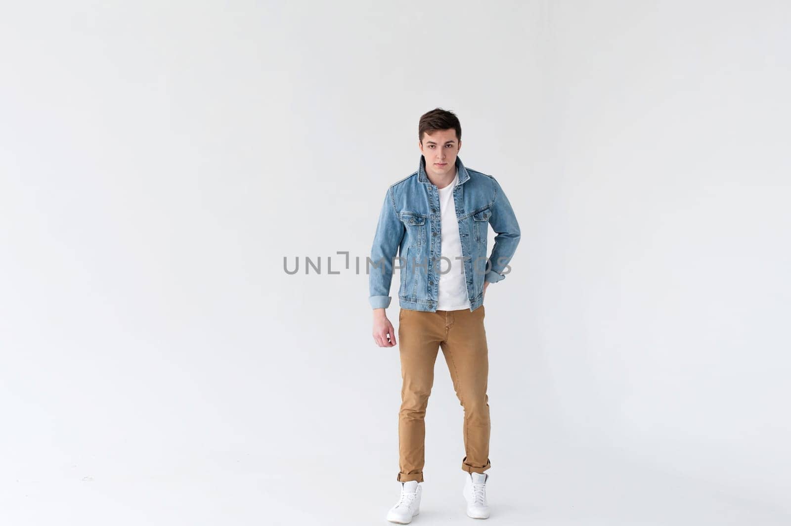Young handsome man of strong Athlete's body, dressed in Blue jeans jacket in style of old school and white t-shirt with brown trousers, Posing on white studio background