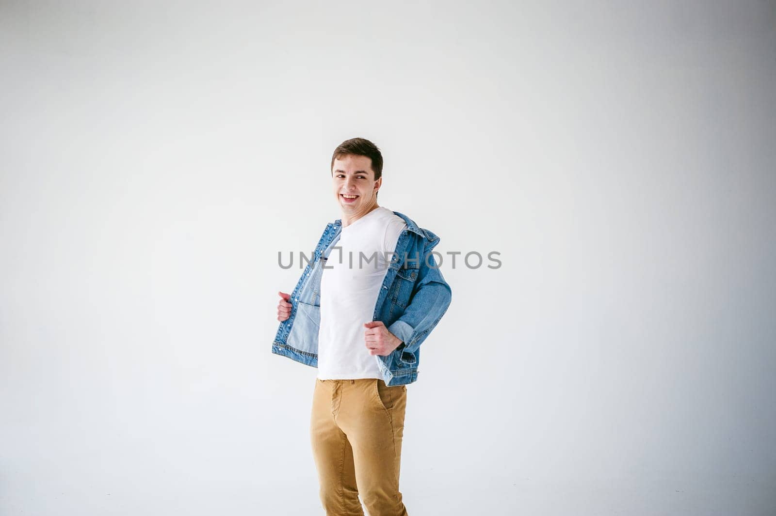Young handsome man of strong Athlete's body, dressed in Blue jeans jacket in style of old school and white t-shirt with brown trousers, Posing on white studio background