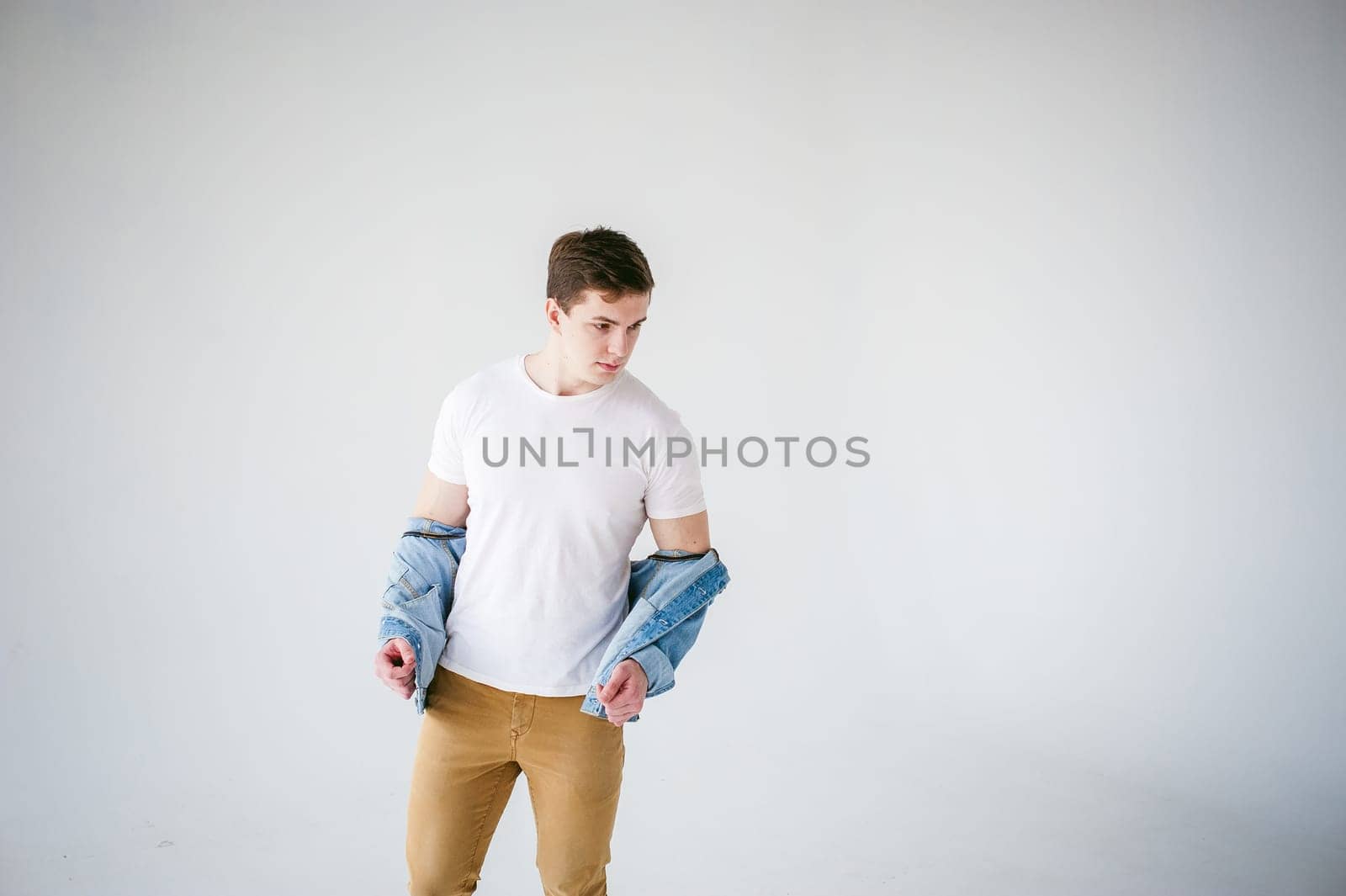 Young handsome man of strong Athlete's body, dressed in Blue jeans jacket in style of old school and white t-shirt with brown trousers, Posing on white studio background