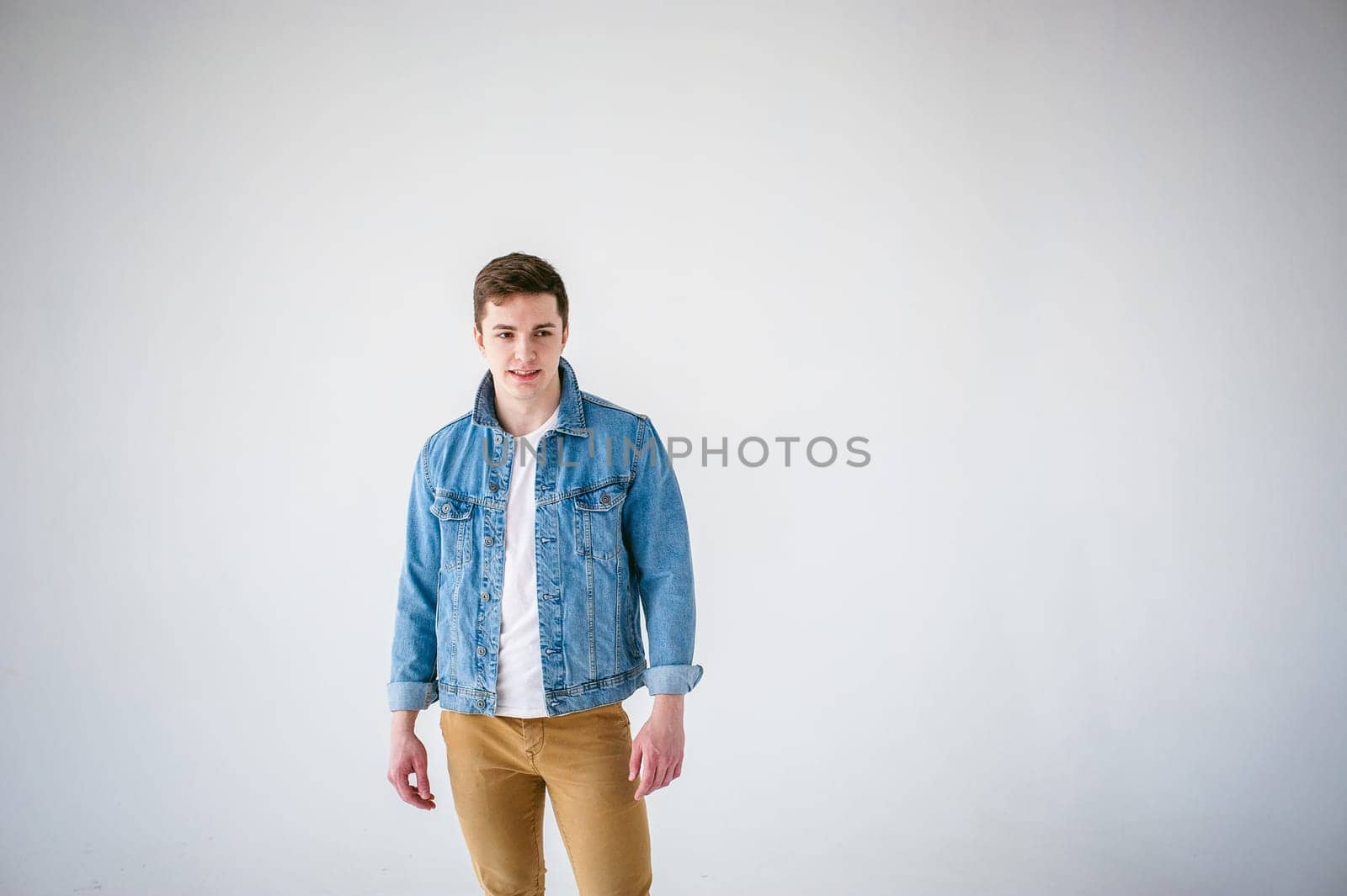 Young handsome man of strong Athlete's body, dressed in Blue jeans jacket in style of old school and white t-shirt with brown trousers, Posing on white studio background