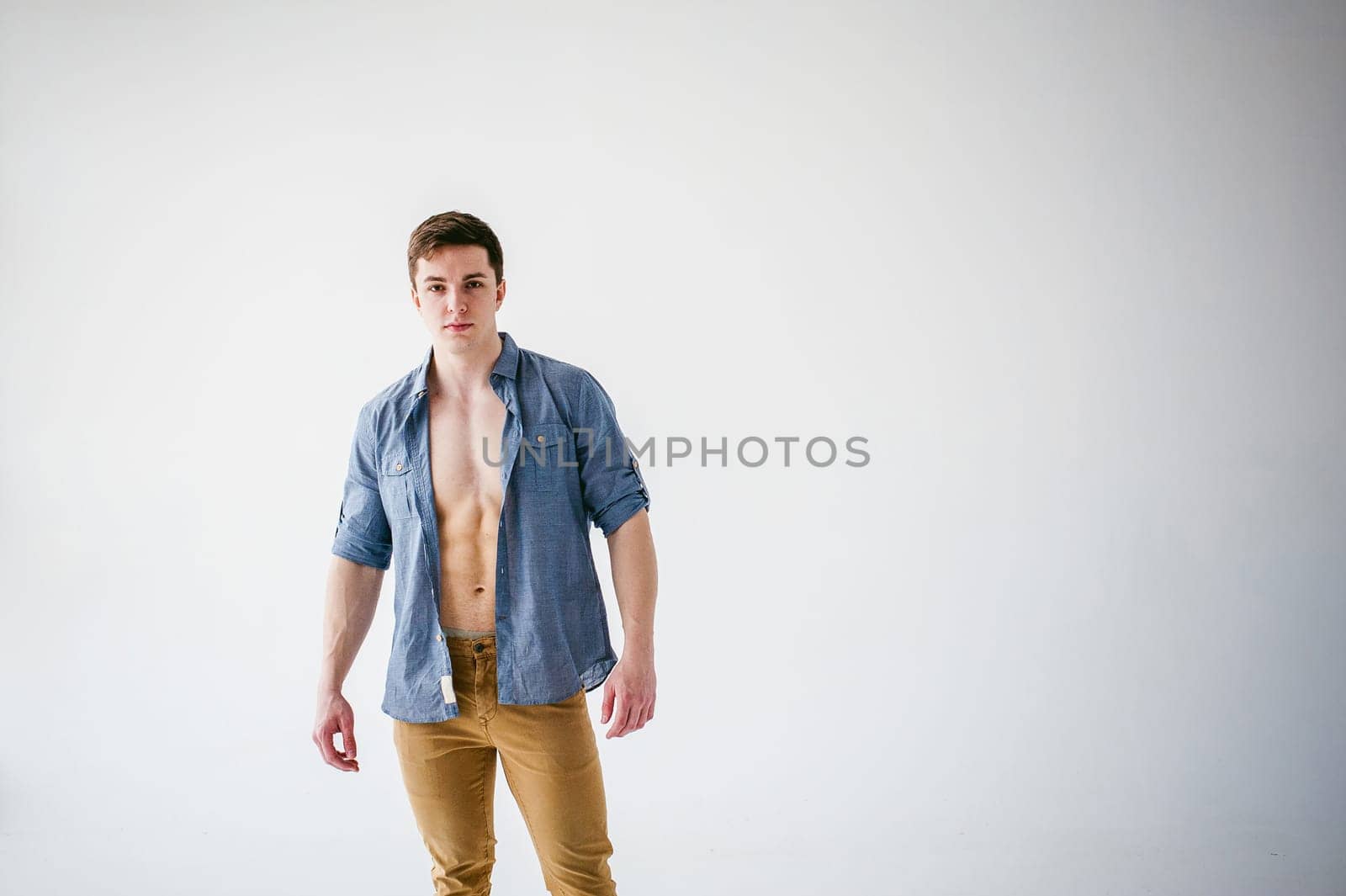 portrait young sexy handsome man of strong Athlete's body with bare torso, Dressed in an unbuttoned shirt and brown trousers, Posing on white studio background