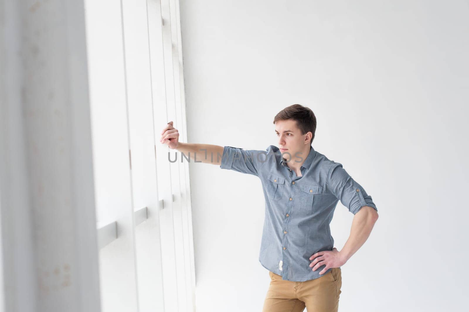 Young handsome man of strong athletic build, dressed in a shirt and brown trousers, Posing on white studio background, rested hand near window in full wall