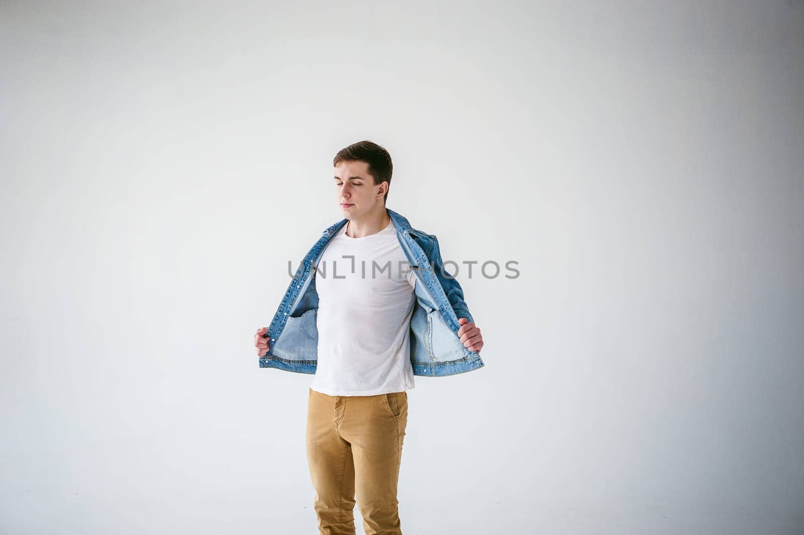 Young handsome man of strong Athlete's body, dressed in Blue jeans jacket in style of old school and white t-shirt with brown trousers, Posing on white studio background