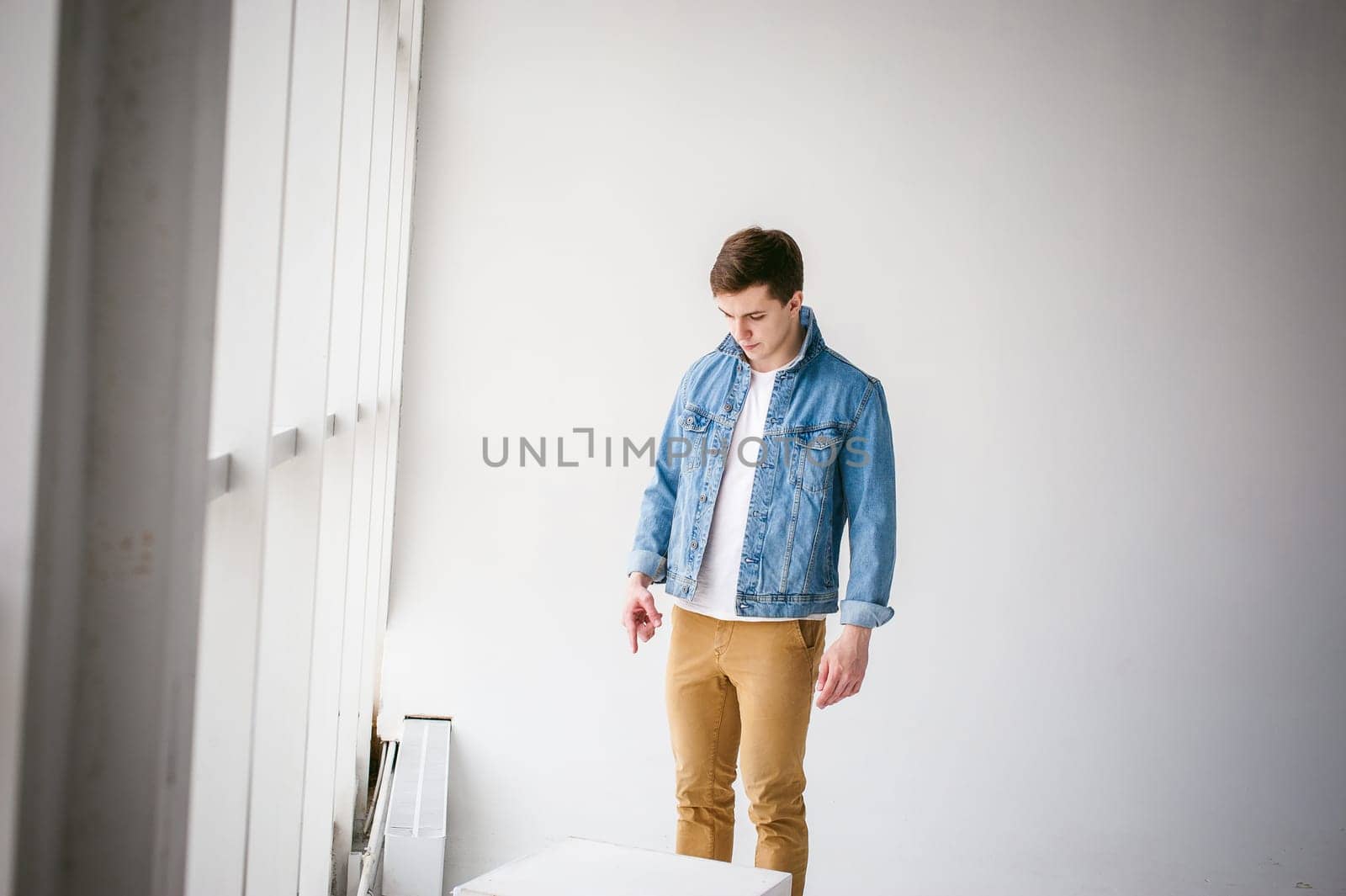 Young handsome man of strong Athlete's body, dressed in Blue jeans jacket in style of old school and white t-shirt with brown trousers, Posing on white studio background, Near a large light window