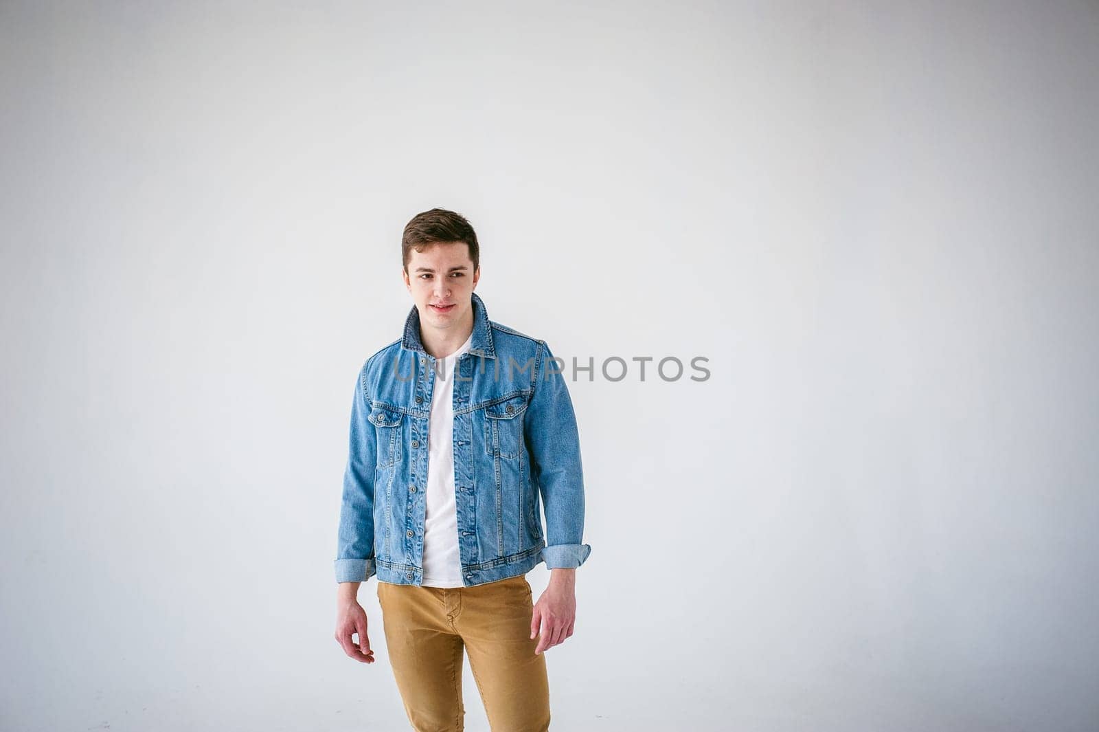 Young handsome man of strong Athlete's body, dressed in Blue jeans jacket in style of old school and white t-shirt with brown trousers, Posing on white studio background