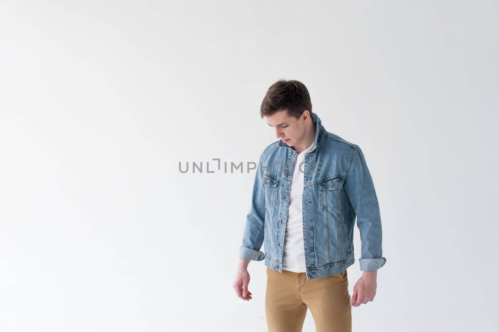Young handsome man of strong Athlete's body, dressed in Blue jeans jacket in style of old school and white t-shirt with brown trousers, Posing on white studio background