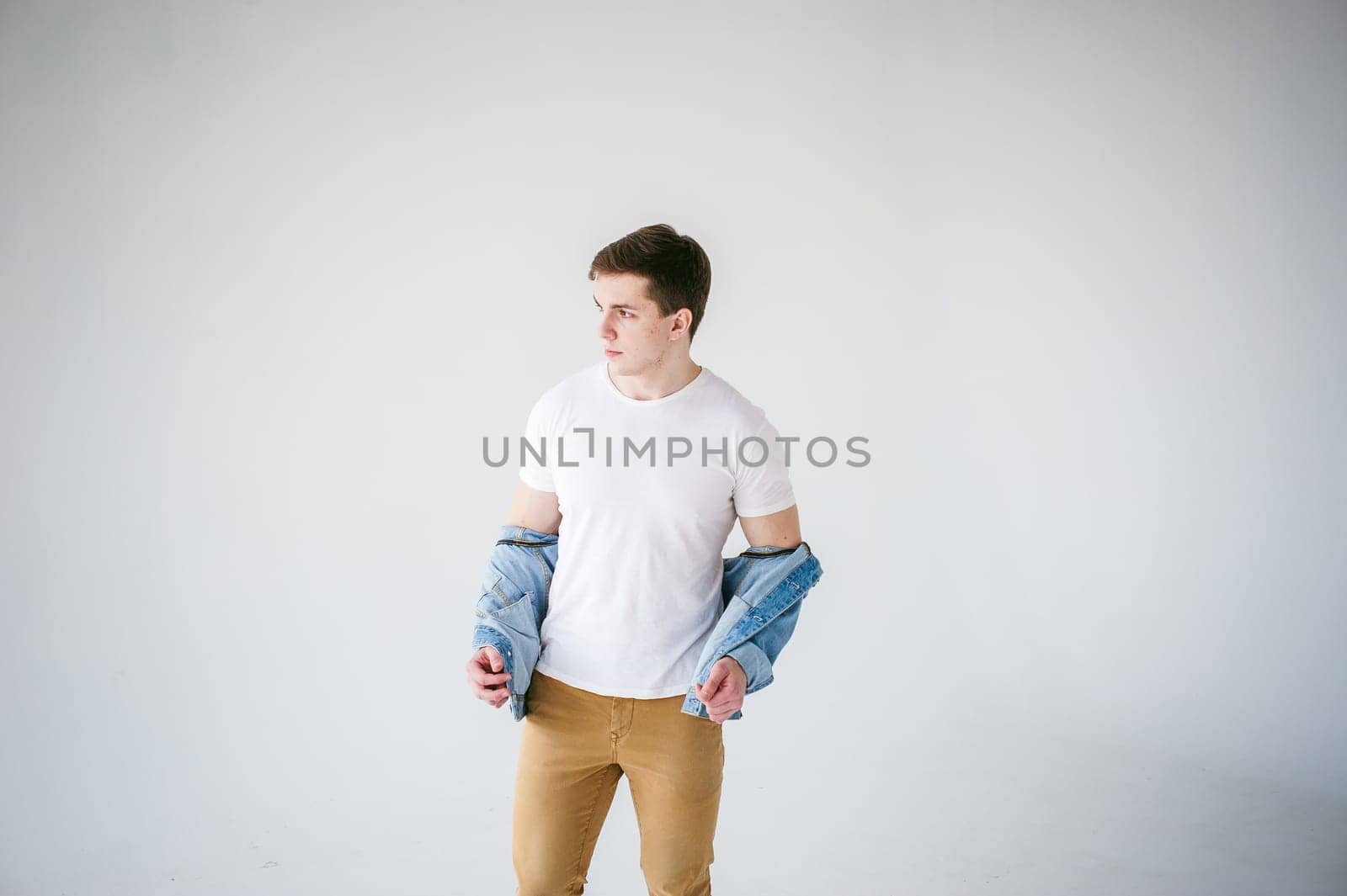 Young handsome man of strong Athlete's body, dressed in Blue jeans jacket in style of old school and white t-shirt with brown trousers, Posing on white studio background