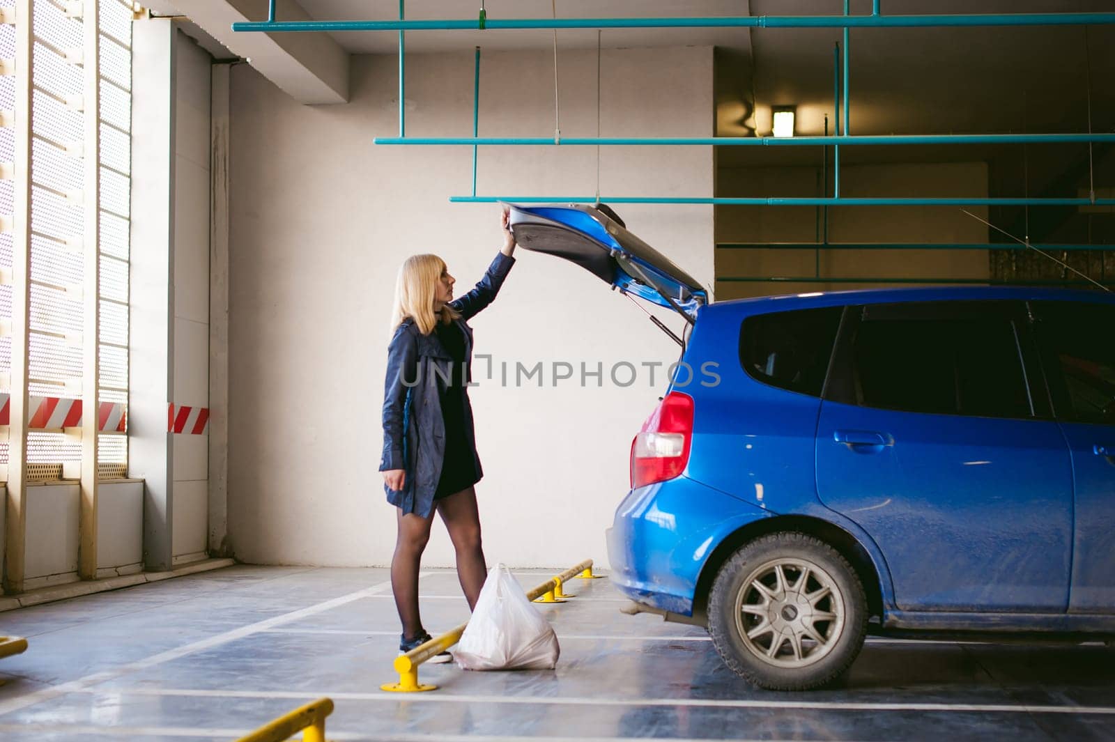 blonde woman in raincoat and pantyhose stacks purchases from grocery store in trunk of the car blue color in covered parking of shopping center