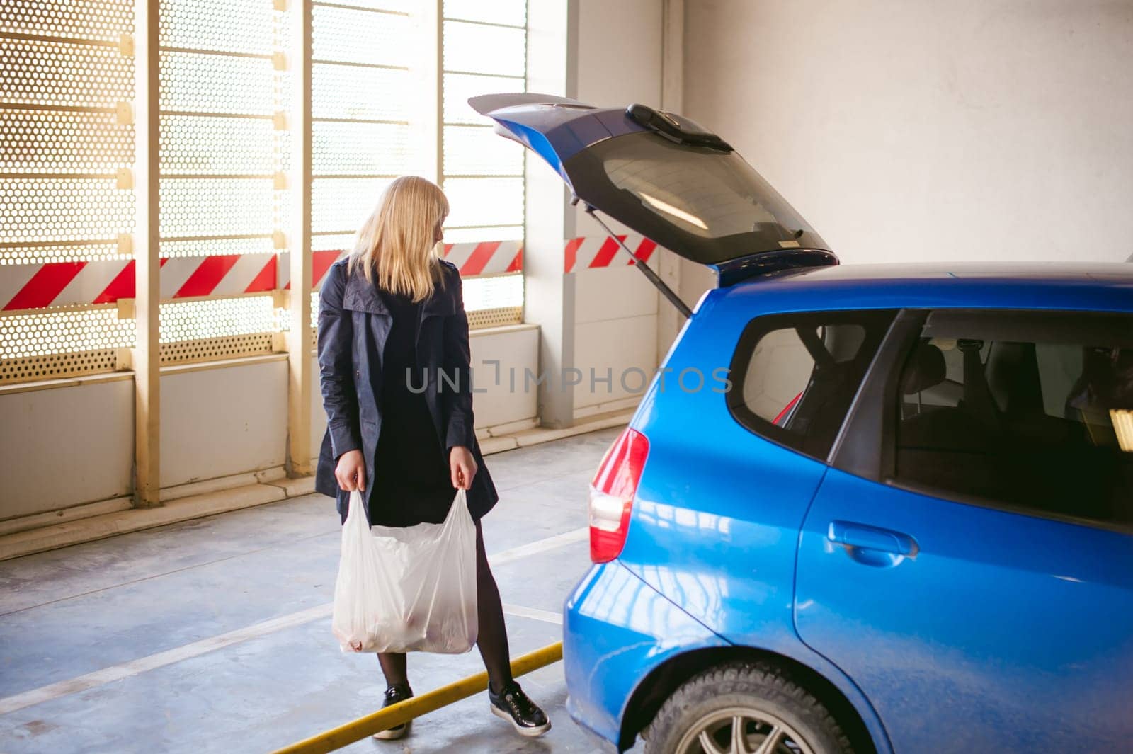 blonde woman in raincoat and pantyhose stacks purchases from grocery store in trunk of the car blue color in covered parking of shopping center