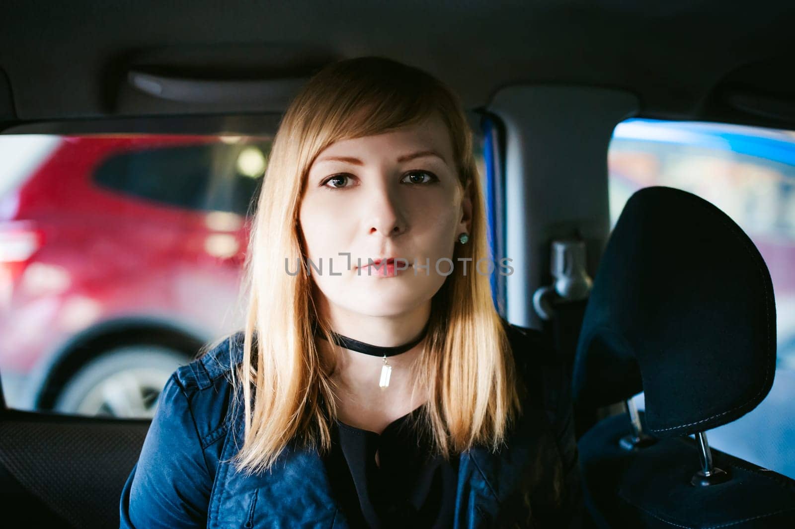 Portrait of a blond woman sitting in a car in the back seat, dressed in a raincoat and a checker on the neck, the picture is taken through the back of the car