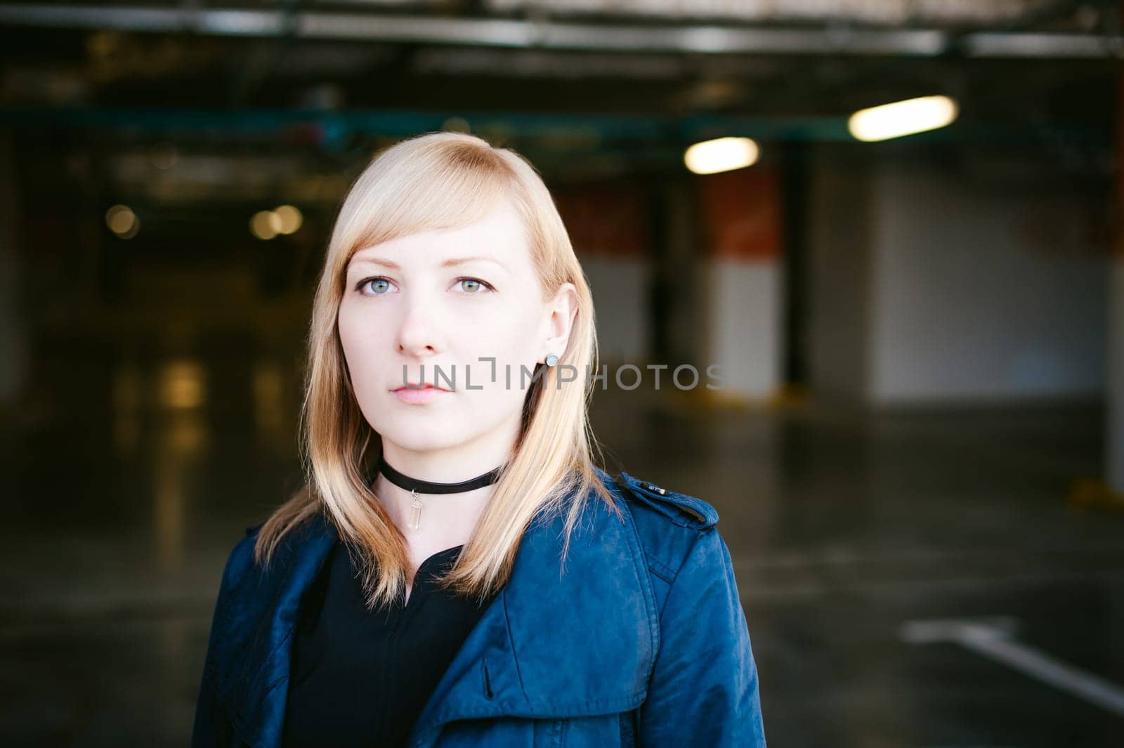 Portrait of lonely blonde woman dressed in dark raincoat with black choker with crystal on her neck, will get bored in covered car park