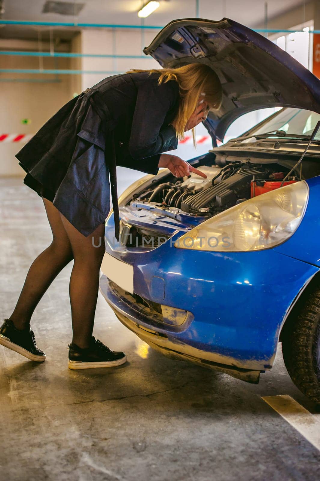 woman near car's hood. young blonde in covered parking of shopping center, stands near car with raised engine compartment hood, solves problem with malfunction by phone call to service