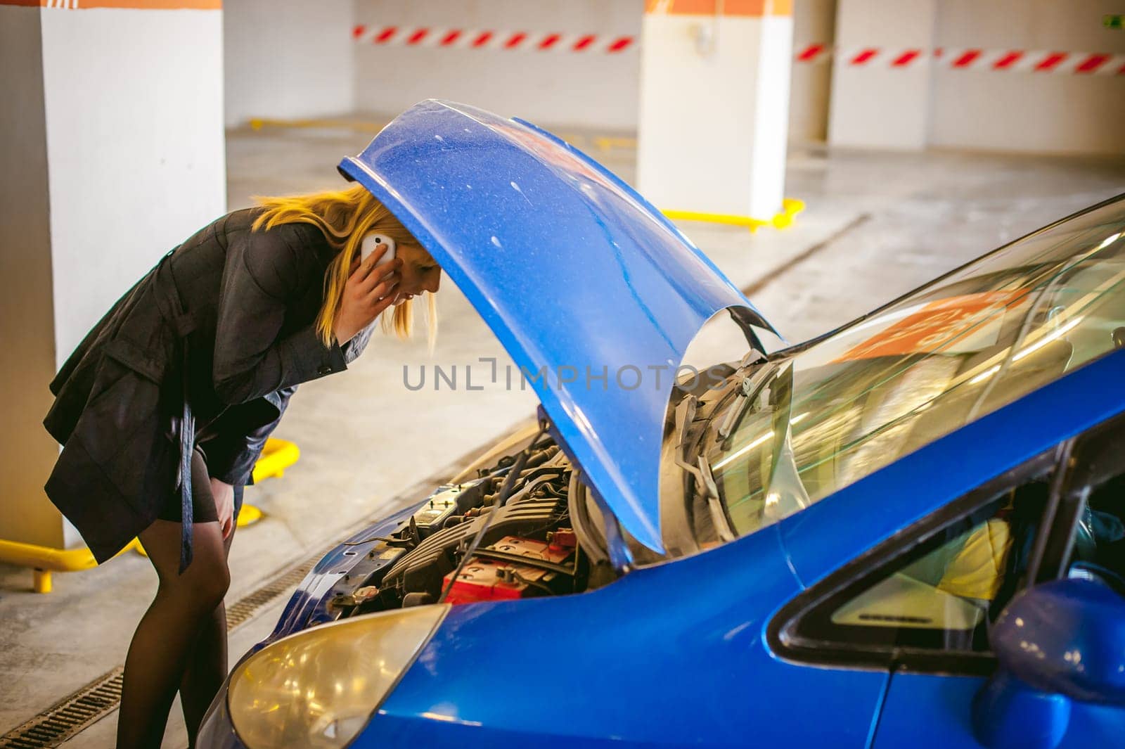 woman near car's hood. young blonde in covered parking of shopping center, stands near car with raised engine compartment hood, solves problem with malfunction by phone call to service