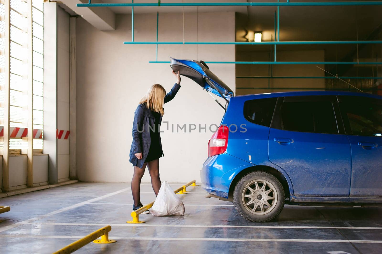 blonde woman in raincoat and pantyhose stacks purchases from grocery store in trunk of the car blue color in covered parking of shopping center