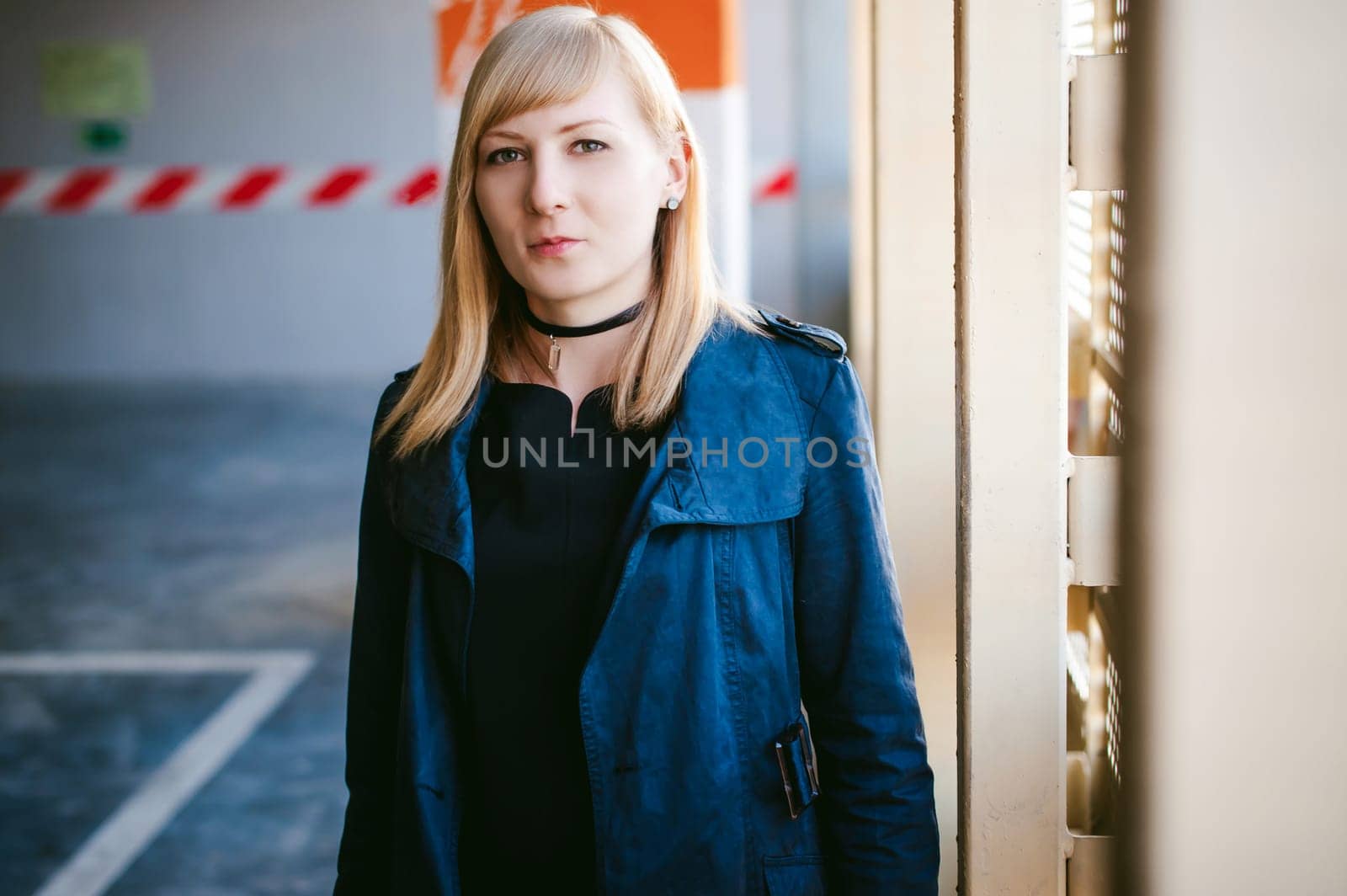 Portrait of lonely blonde woman dressed in dark raincoat with black choker with crystal on her neck, will get bored in covered car park
