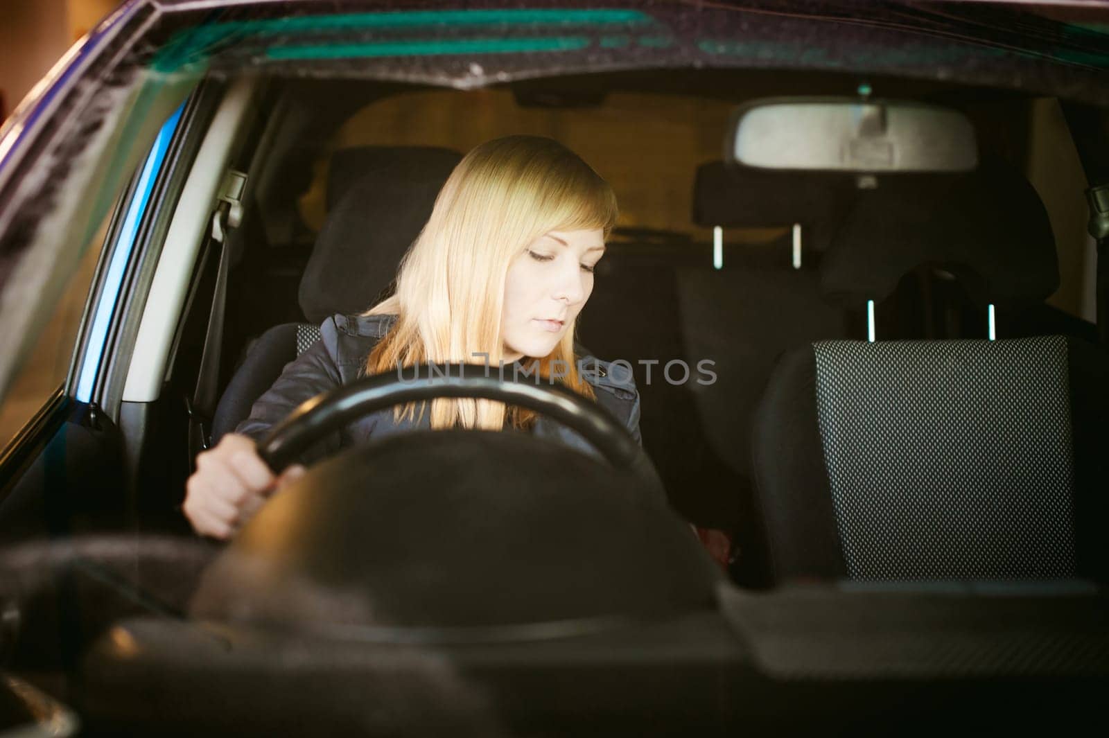 Portrait blonde woman sitting behind wheel in driver's seat of blue car. Hands on handlebars, driving problems of female