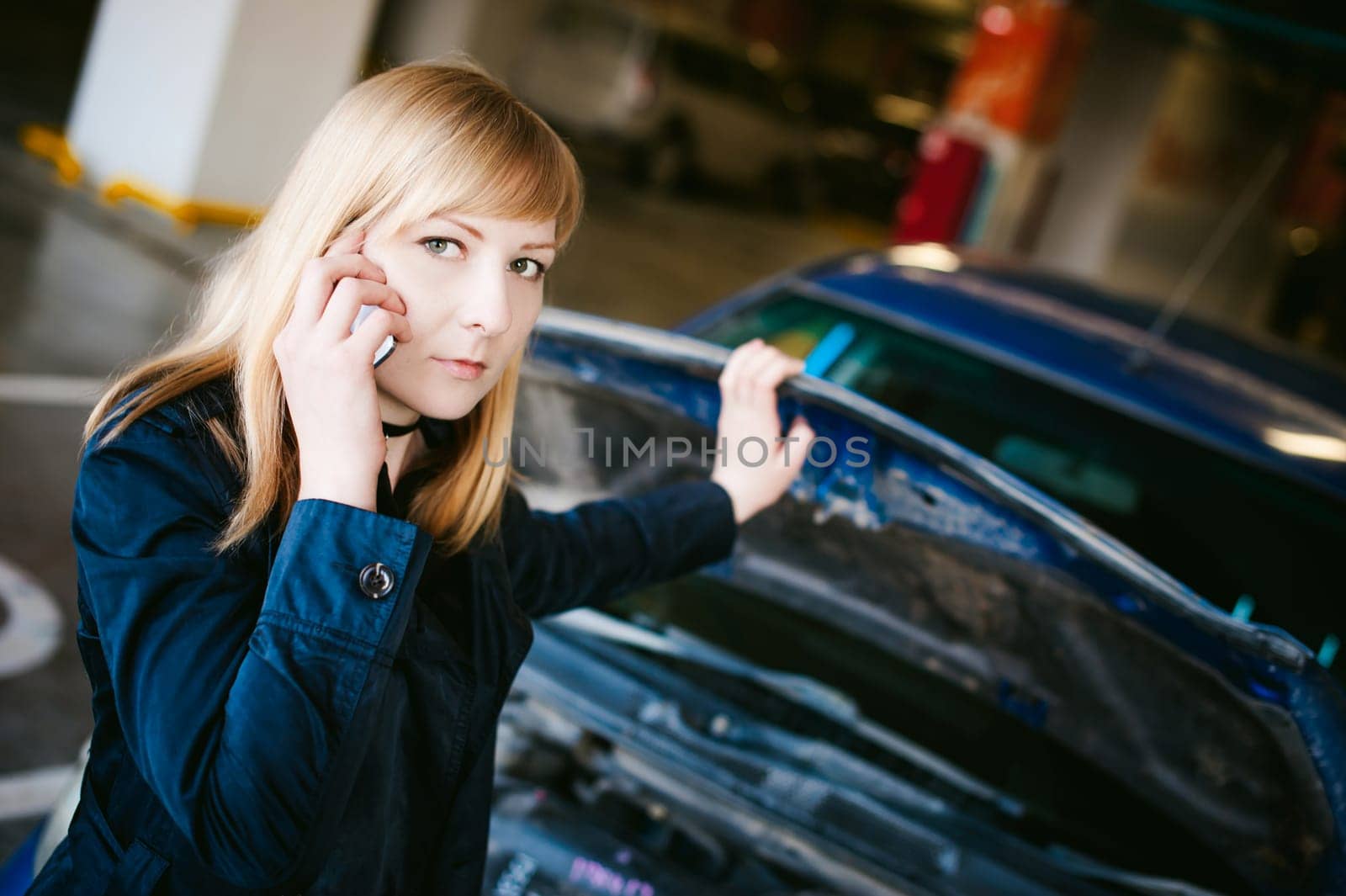 Portrait of blonde woman in black cloak calling phone in car service for help, lifting hood of car and looking at engine, malfunction, diagnosis, service