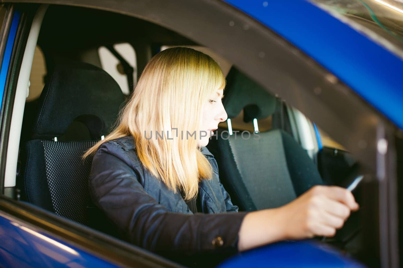 Portrait blonde woman sitting behind wheel in driver's seat of blue car. Hands on handlebars, driving problems of female