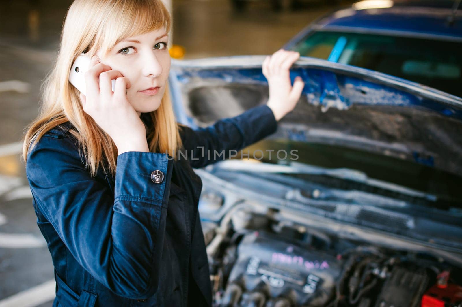 Portrait of blonde woman in black cloak calling phone in car service for help, lifting hood of car and looking at engine, malfunction, diagnosis, service