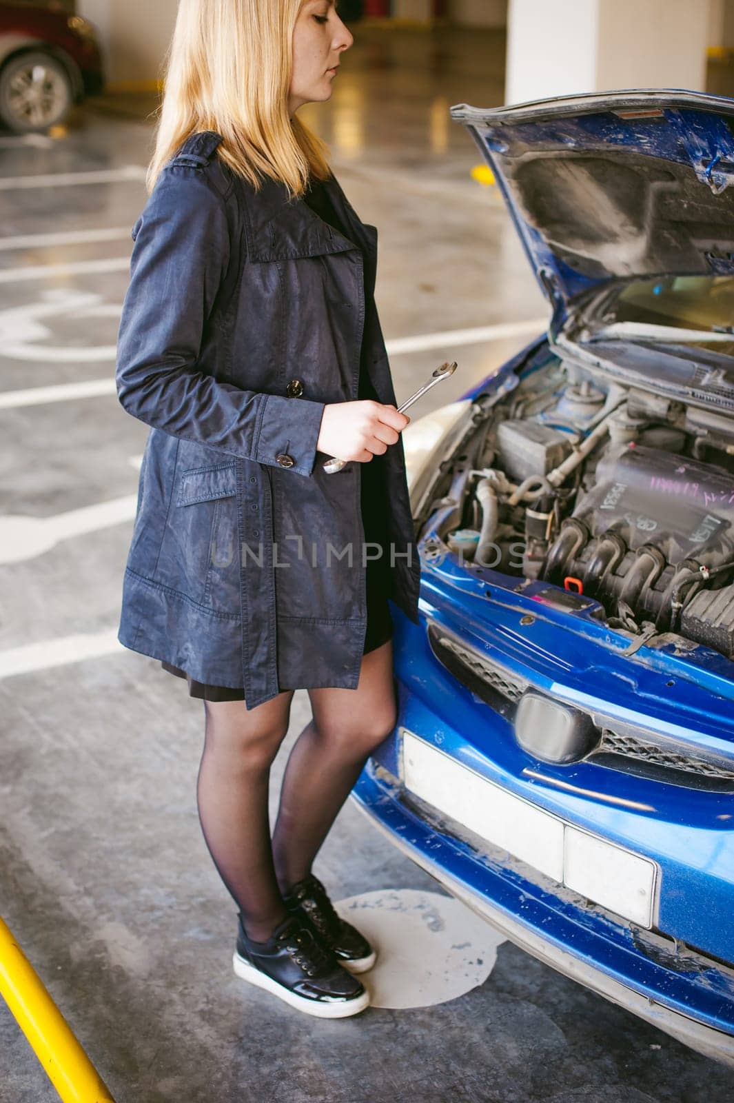 woman near car's hood. young blonde in covered parking of shopping center, stands near car with engine compartment bonnet raised, solves problem with engine malfunction, using carabiner wrench tool