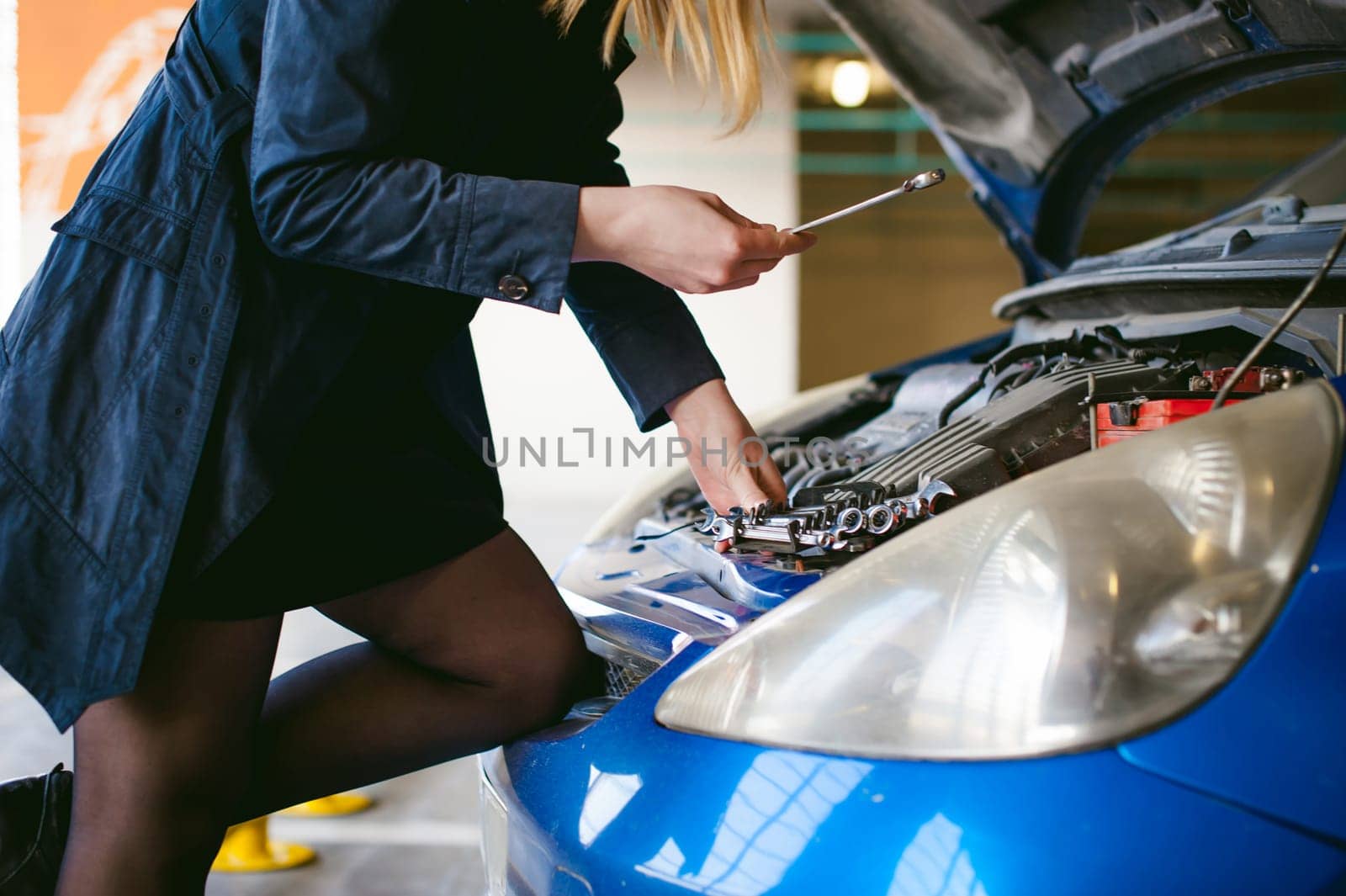 woman near car's hood. young blonde in covered parking of shopping center, stands near car with engine compartment bonnet raised, solves problem with engine malfunction, using carabiner wrench tool