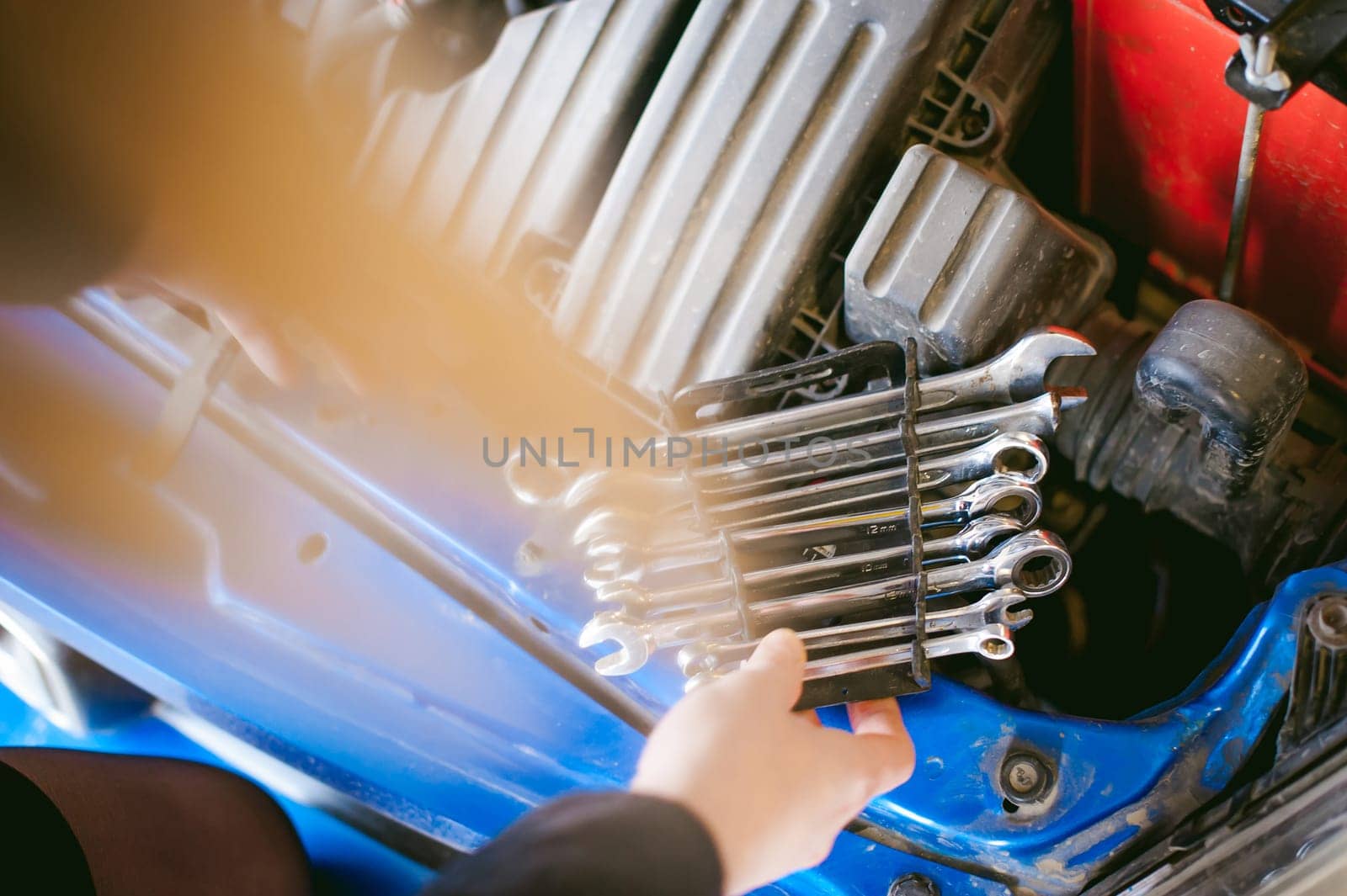 woman near car's hood. young blonde in covered parking of shopping center, stands near car with engine compartment bonnet raised, solves problem with engine malfunction, using carabiner wrench tool