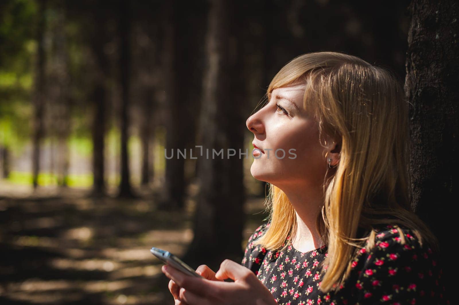 blonde woman dressed summer dress, black pantyhose and sneakers with white sole, resting nature enjoying warm sunny day. Sits under tree alone, holding in hand cell phone, Emotion online communication