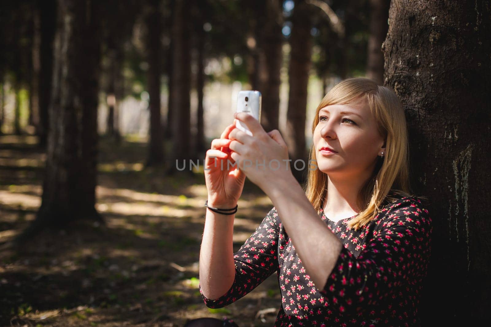 blonde woman dressed summer dress, black pantyhose and sneakers with white sole, resting nature enjoying warm sunny day. Sits under tree alone, holding in hand cell phone, Emotion online communication