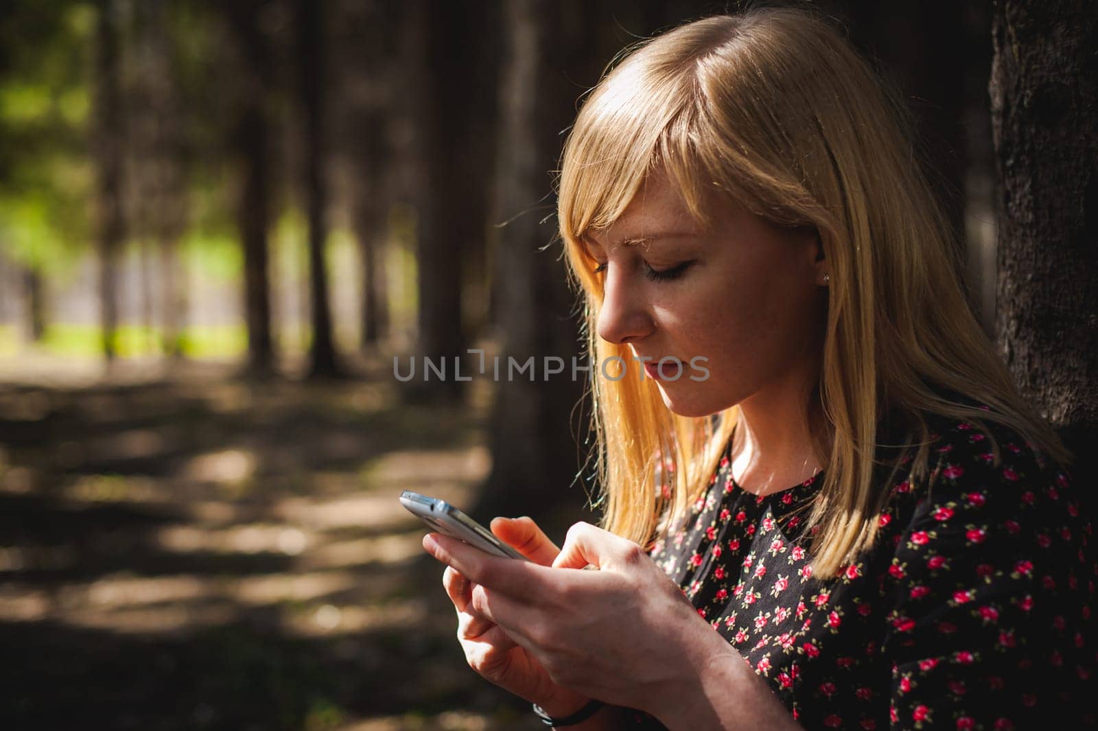 blonde woman dressed summer dress, black pantyhose and sneakers with white sole, resting nature enjoying warm sunny day. Sits under tree alone, holding in hand cell phone, Emotion online communication