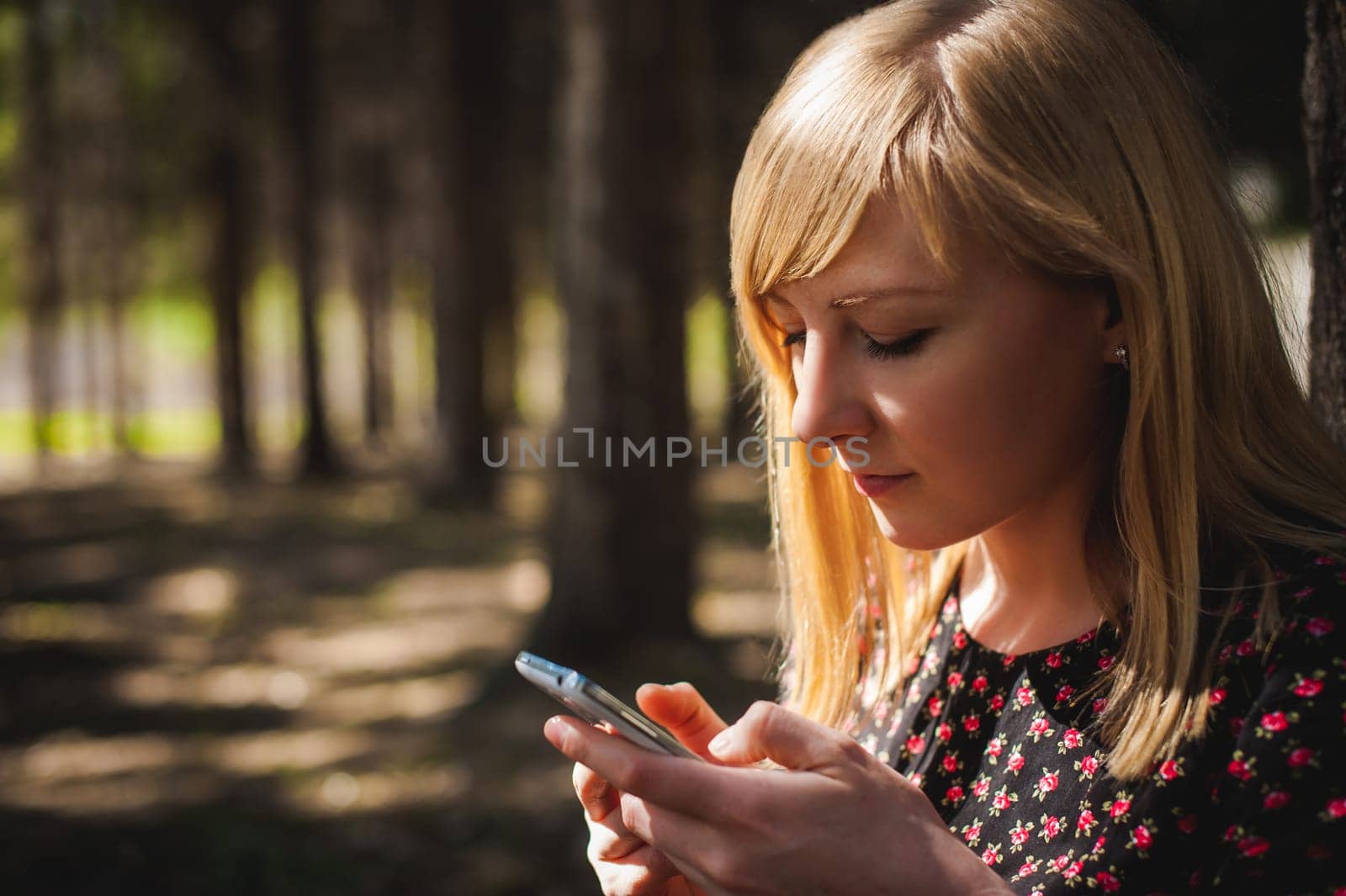 blonde woman dressed summer dress, black pantyhose and sneakers with white sole, resting nature enjoying warm sunny day. Sits under tree alone, holding in hand cell phone, Emotion online communication