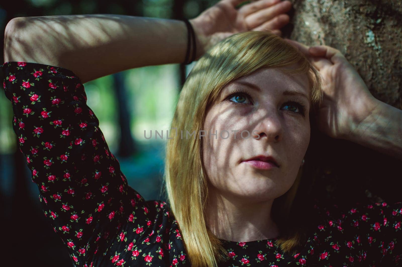 Close-up portrait of blonde woman Hands at head dressed in summer dress. Girl in woods against background of coniferous trees in open air, natural beauty, sunlight shadows on face from branches
