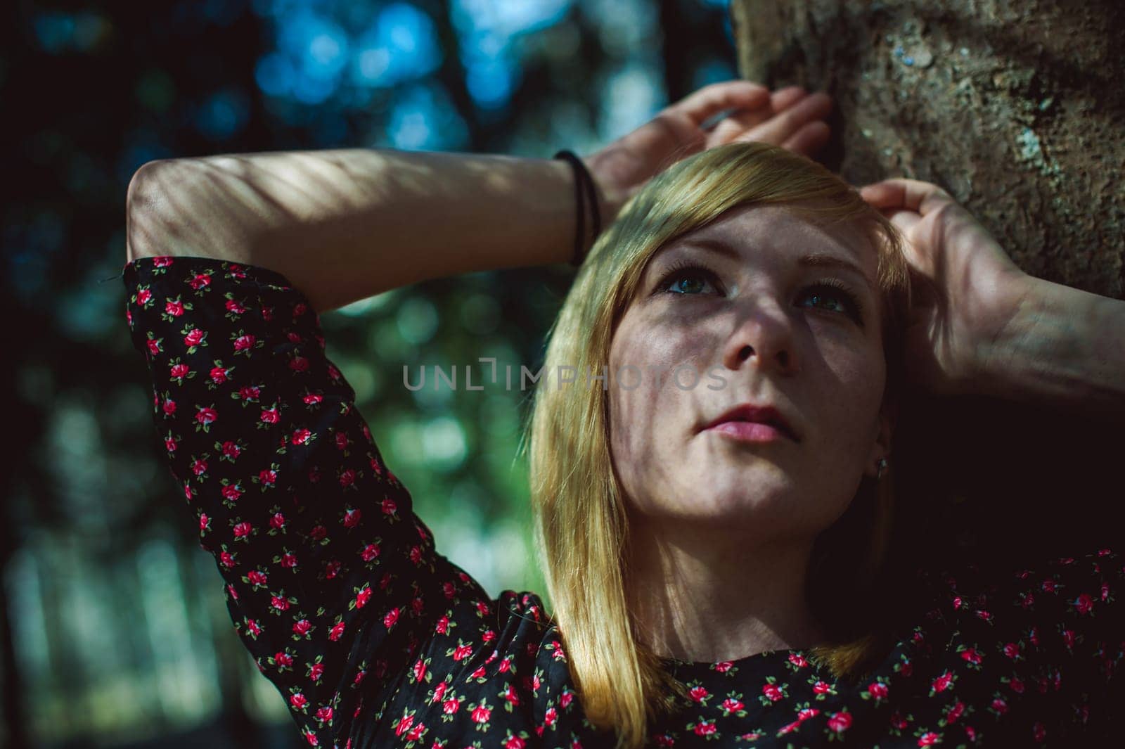Close-up portrait of blonde woman Hands at head dressed in summer dress. Girl in woods against background of coniferous trees in open air, natural beauty, sunlight shadows on face from branches