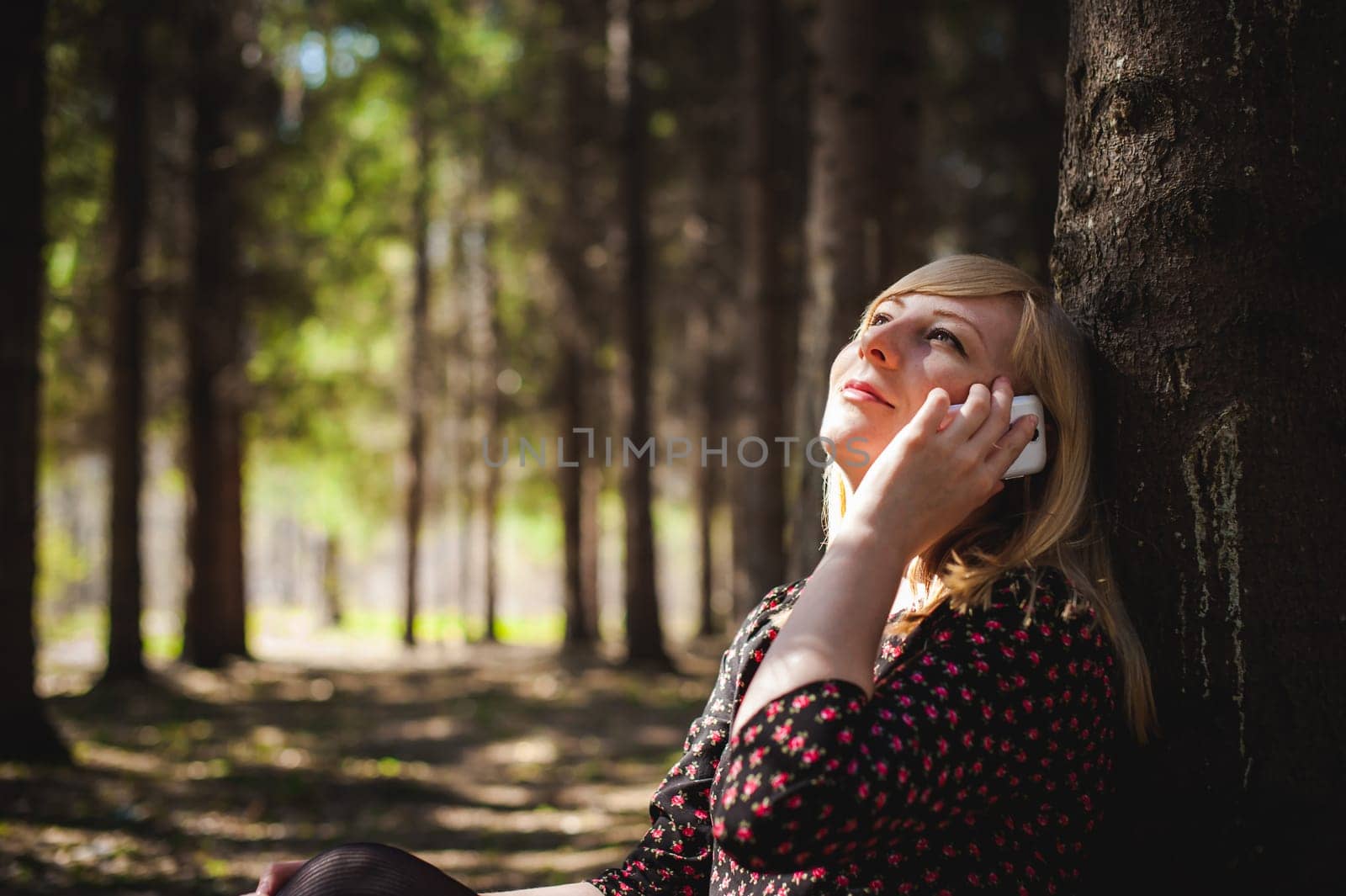 Walk girl coniferous forest park. blonde woman dressed summer dress, black pantyhose and sneakers with white sole, resting nature enjoying warm sunny day. Sits under tree alone, Speaks cell phone