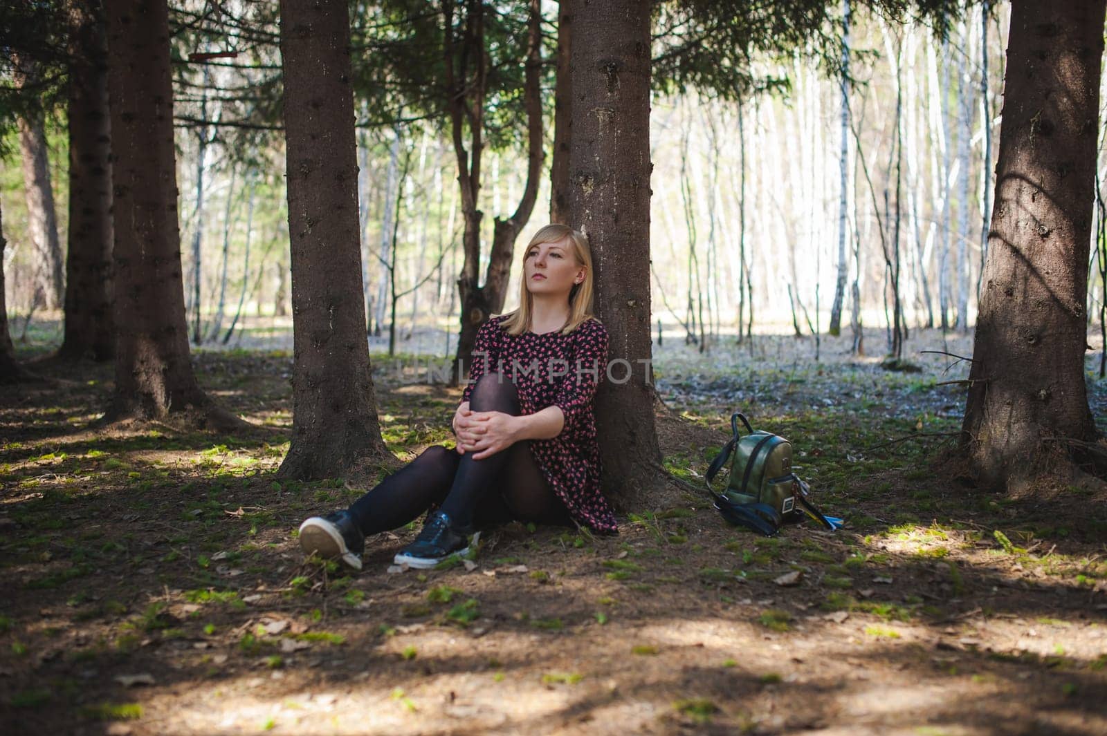 Walk girl coniferous forest park. blond woman with rucksack dressed summer dress, black pantyhose and sneakers with white sole, resting nature enjoying warm sunny afternoon. Sits under tree alone