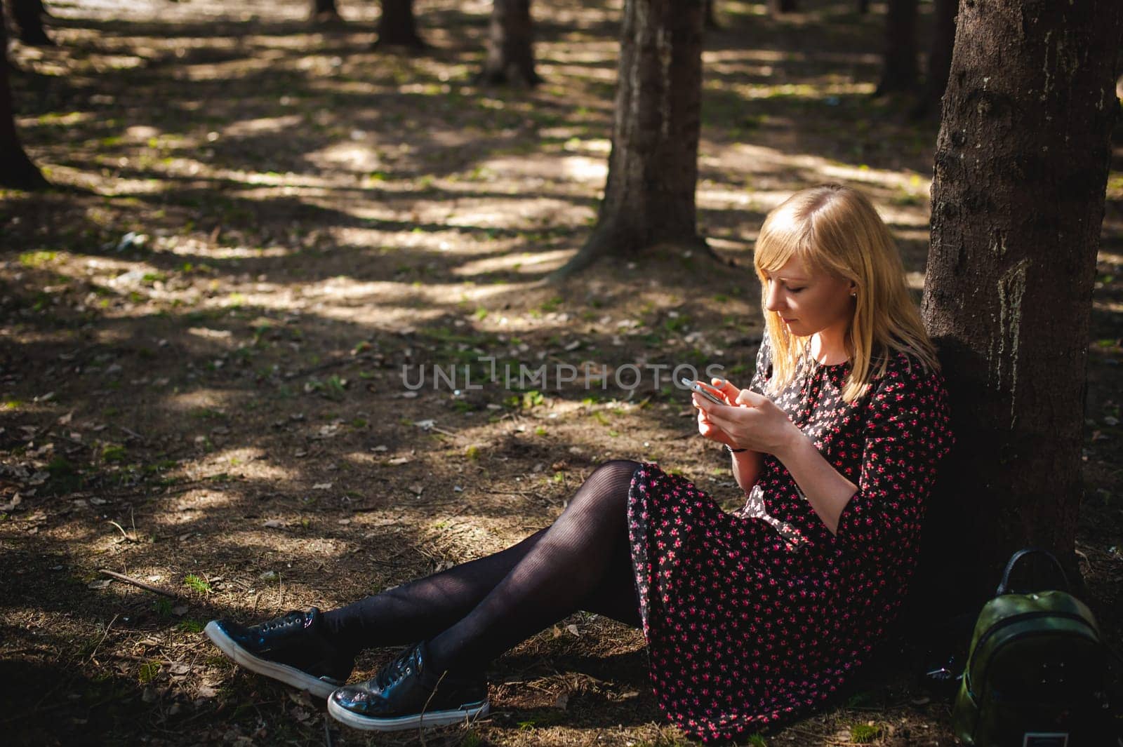 blonde woman dressed summer dress, black pantyhose and sneakers with white sole, resting nature enjoying warm sunny day. Sits under tree alone, holding in hand cell phone, Emotion online communication