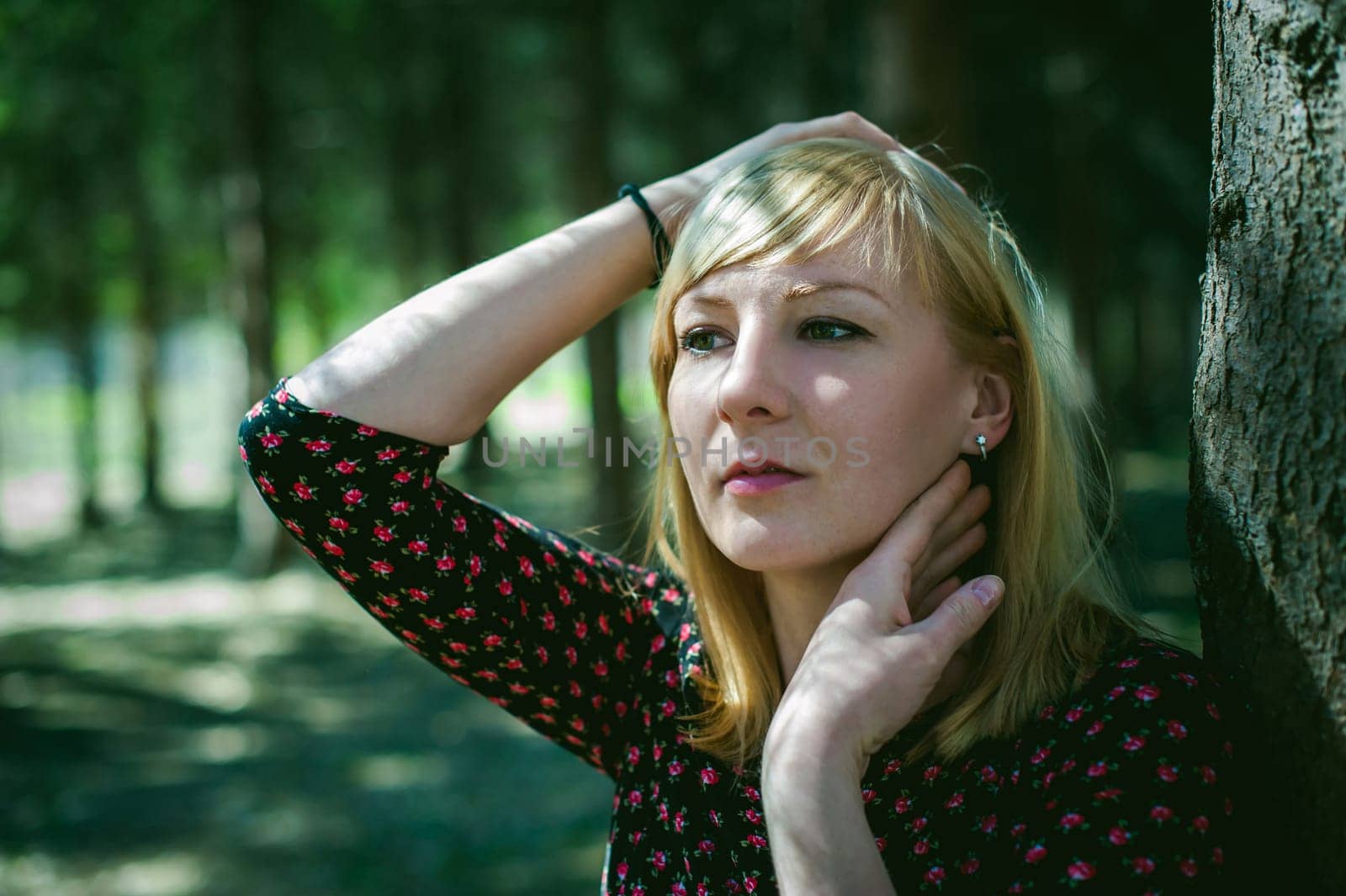 Close-up portrait of blonde woman Hands at head dressed in summer dress. Girl in woods against background of coniferous trees in open air, natural beauty, sunlight shadows on face from branches
