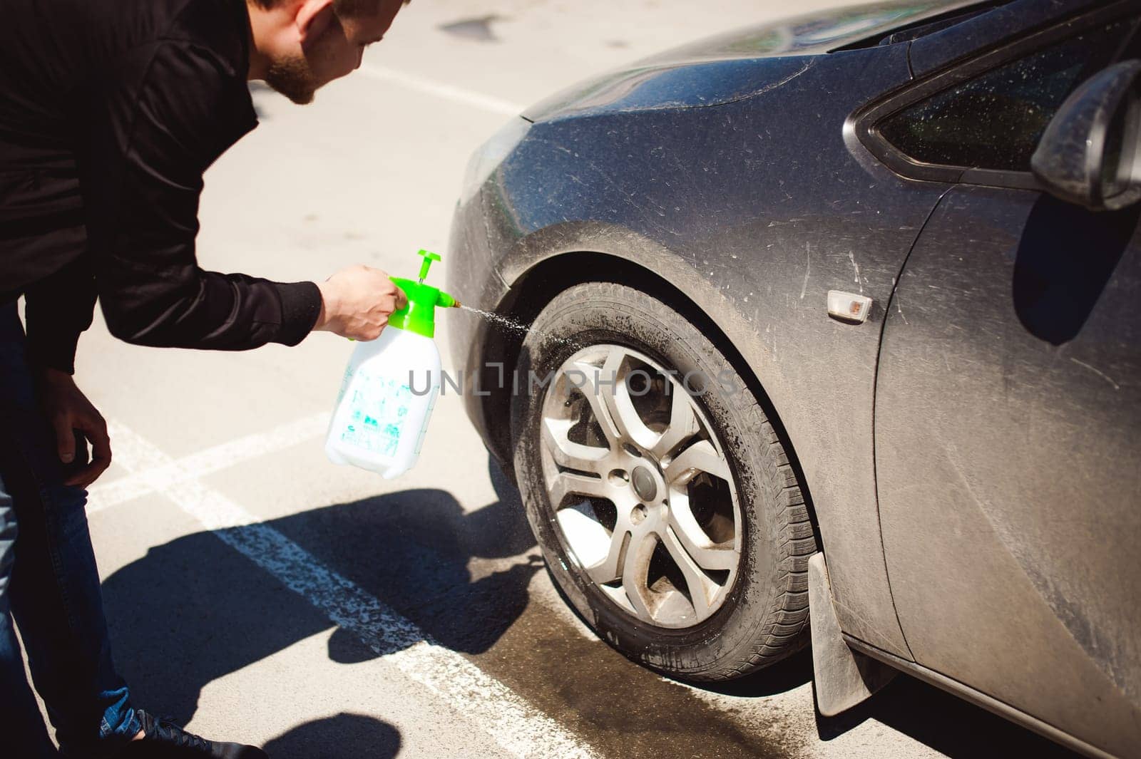young bearded man washes his car's wheel rims, spraying water from spray, in street parking