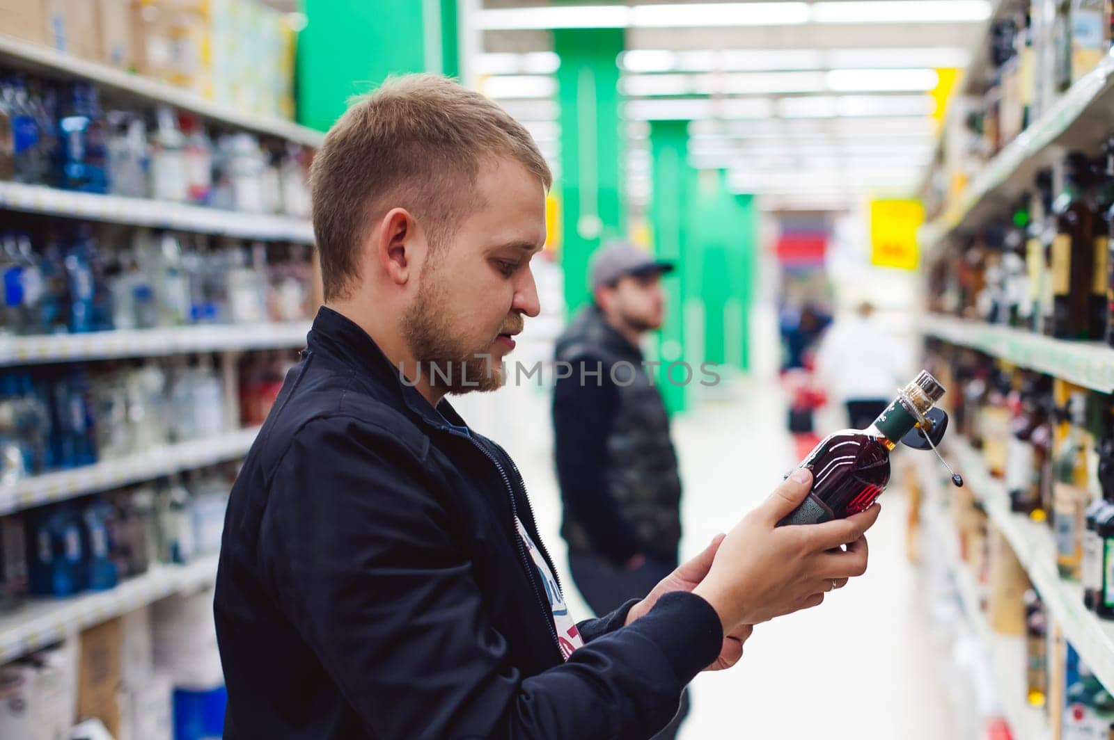 bearded man, In a black jacket, stopped to make my choice in store window, holds a bottle of strong alcohol in his hand, taking from shelf