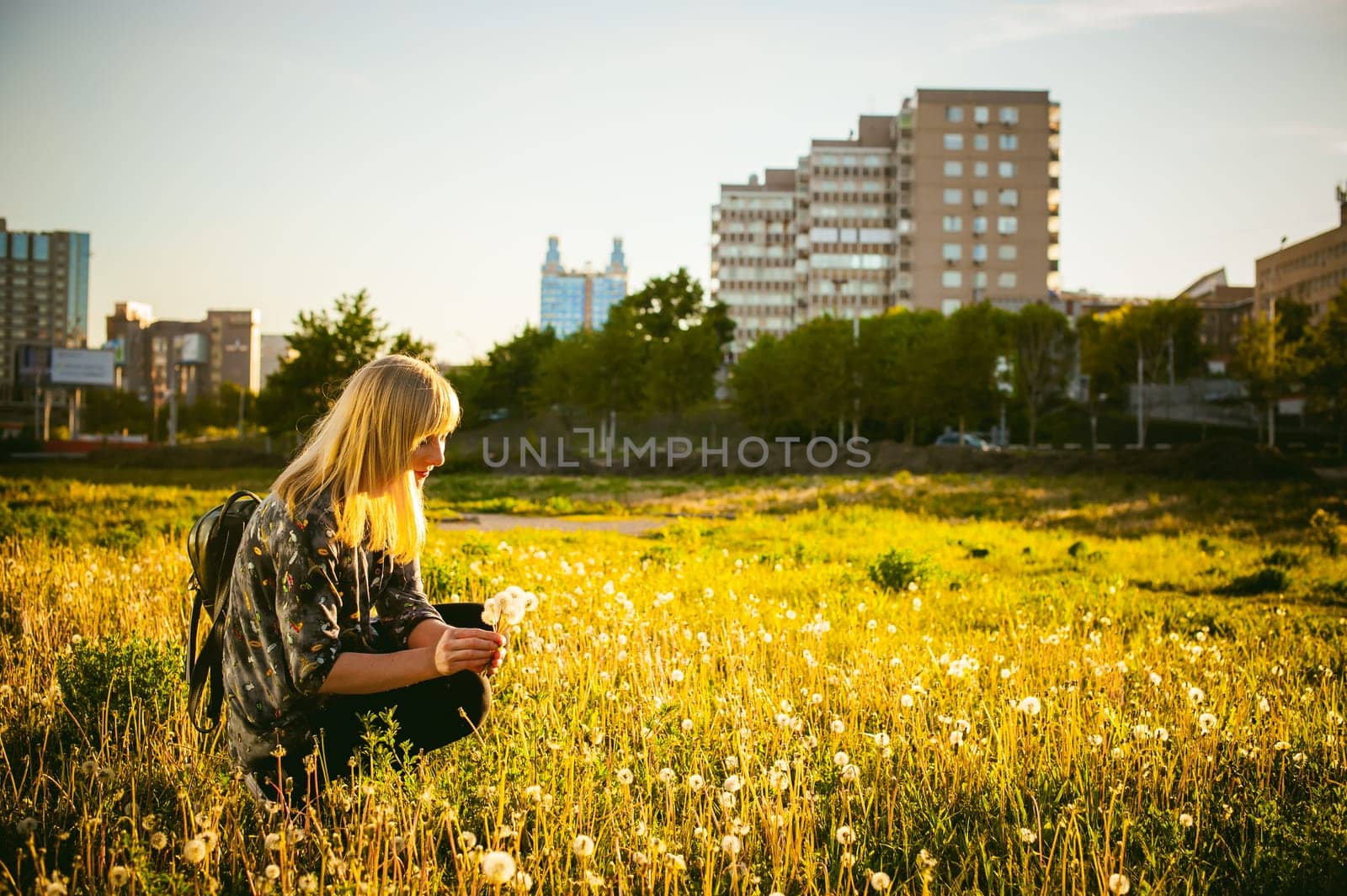 Young blonde girl in dress with shoulder bag, walking on dandelion field, against background of urban architecture, breathing relaxing in cityscape