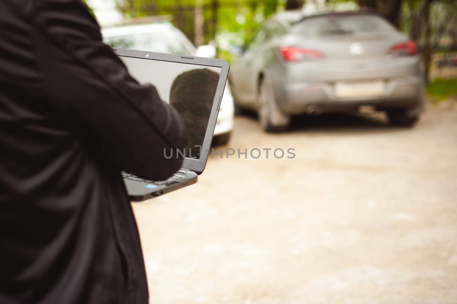 man with a laptop in parking lot in yard near car is doing manipulations with cyber system, concept