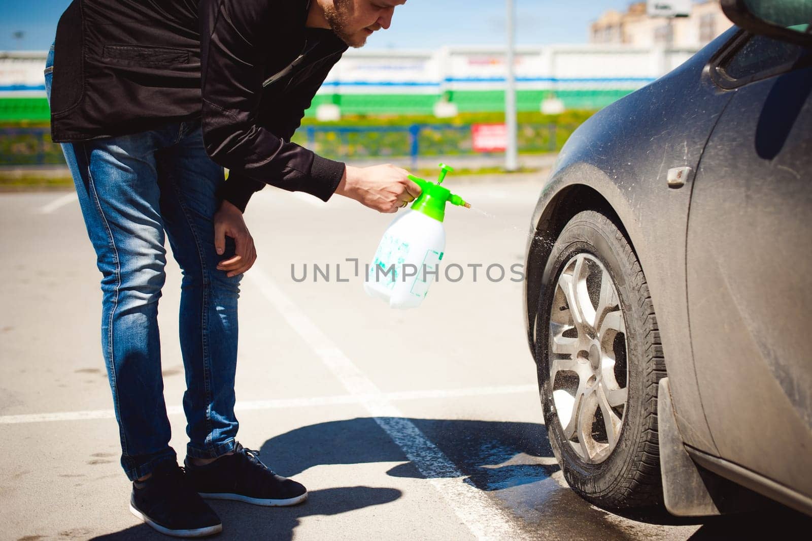 young bearded man washes his car's wheel rims, spraying water from spray, in street parking