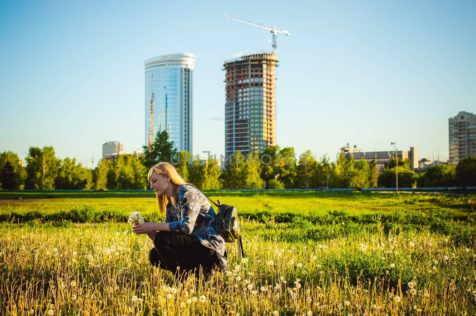 Young blonde girl in dress with shoulder bag, walking on dandelion field, against background of urban architecture, breathing relaxing in cityscape