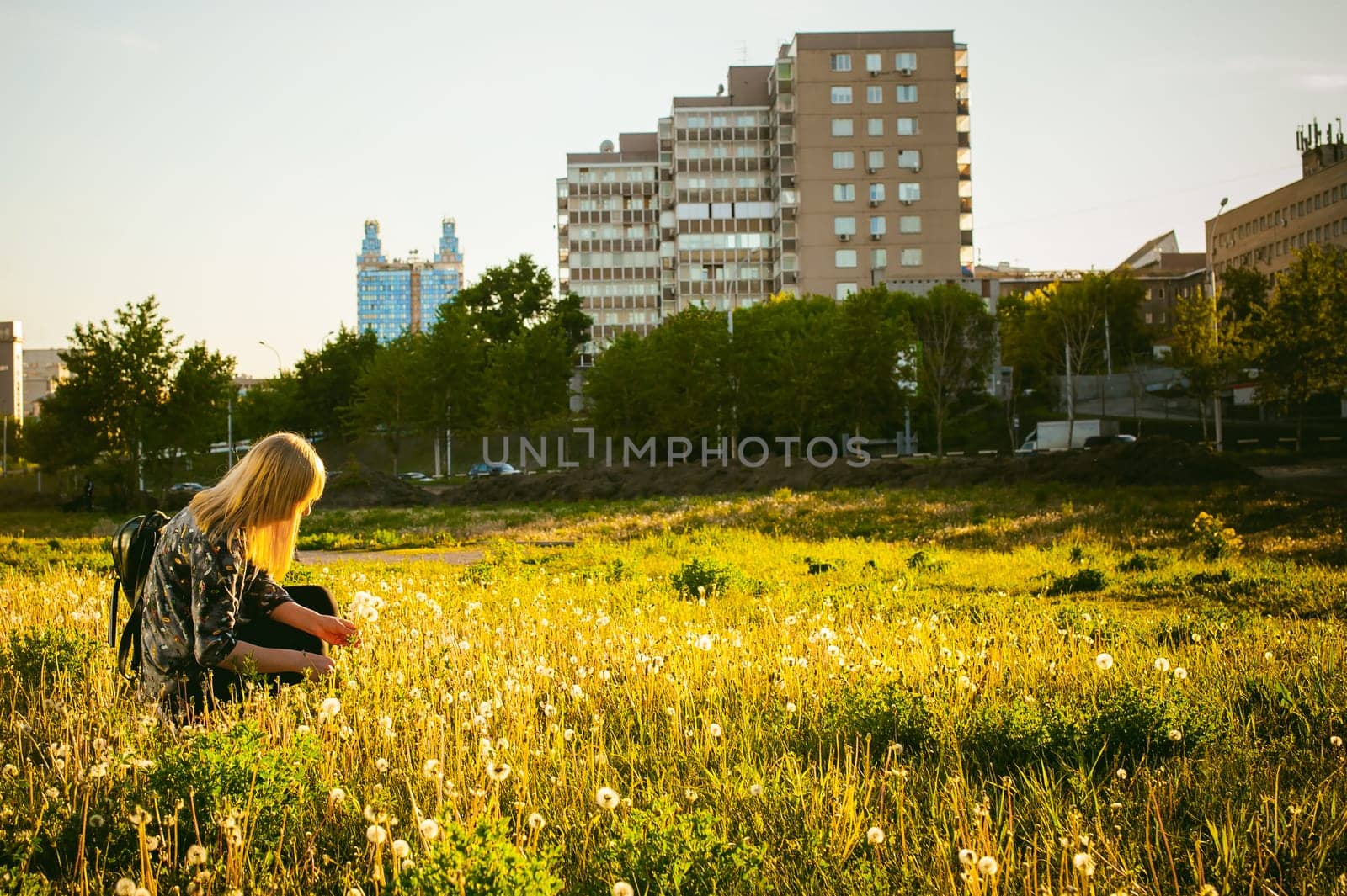 Young blonde girl in dress with shoulder bag, walking on dandelion field, against background of urban architecture, breathing relaxing in cityscape