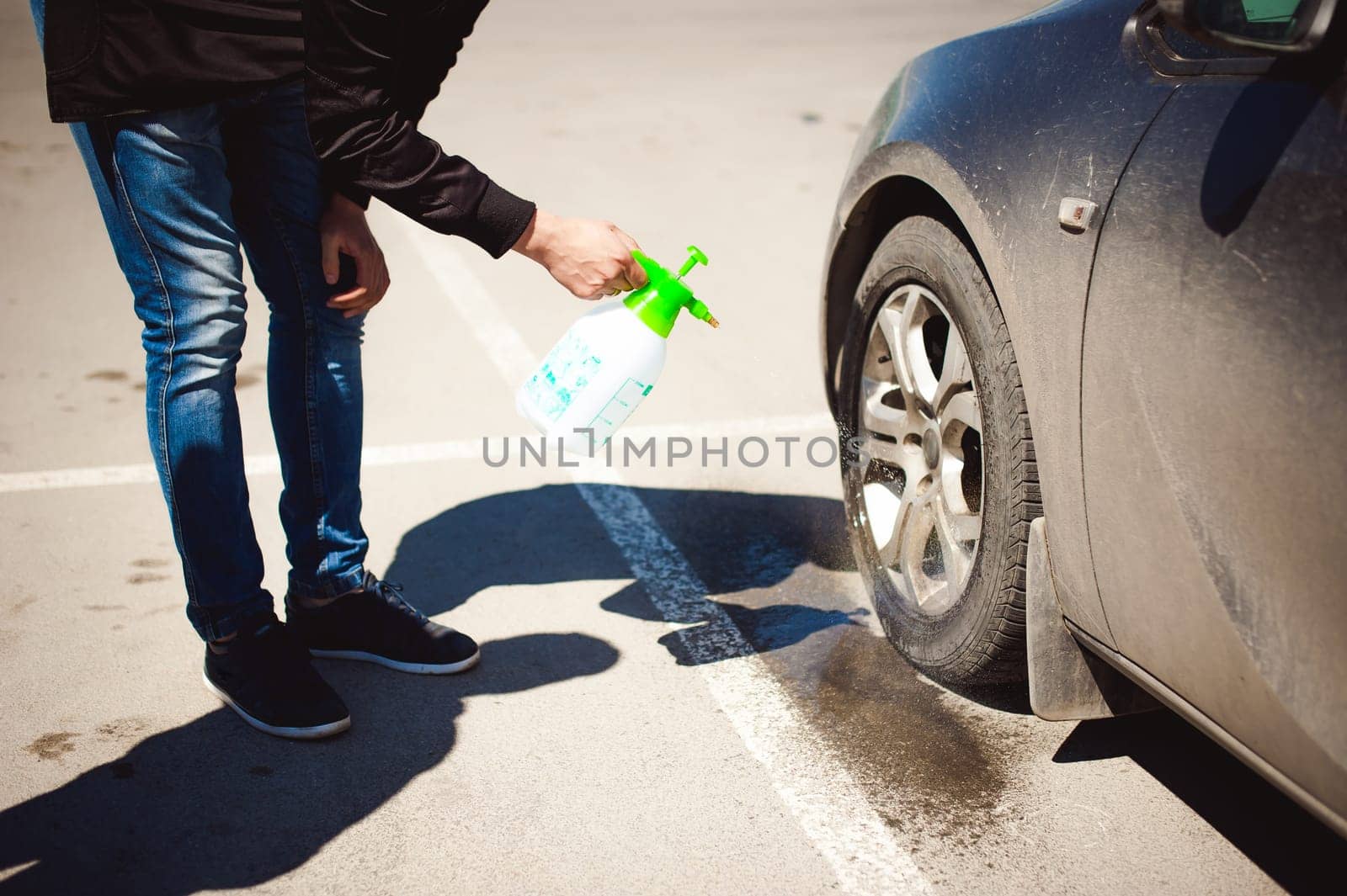young bearded man washes his car's wheel rims, spraying water from spray, in street parking