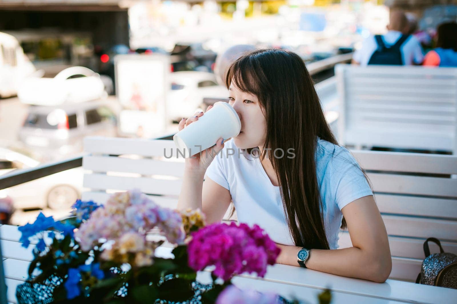 Cute asian young woman in summer cafe outdoors. girl In white T-shirt, with long hair in simple light cozy interior of restaurant Urban style, drinking coffee drink, sitting at table with flowers