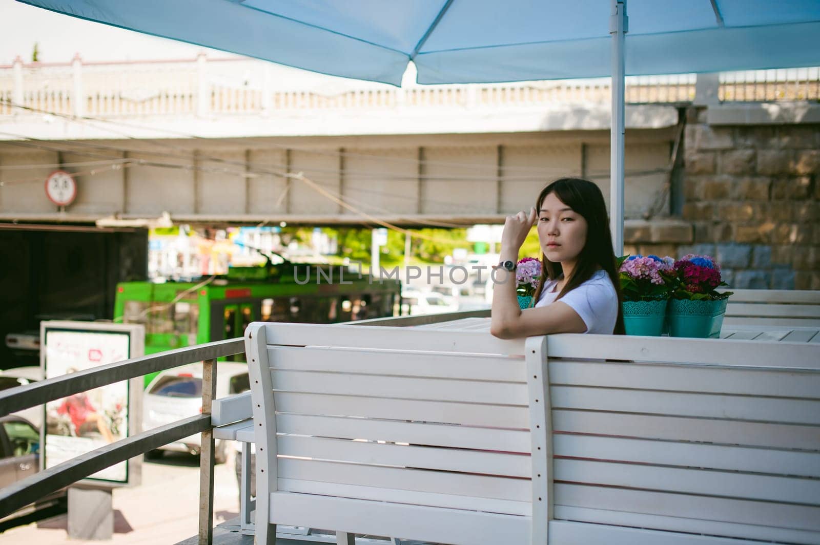 Cute asian young woman in summer cafe outdoors. girl In white T-shirt, with long hair in simple light cozy interior of restaurant Urban style, drinking coffee drink, sitting at table with flowers