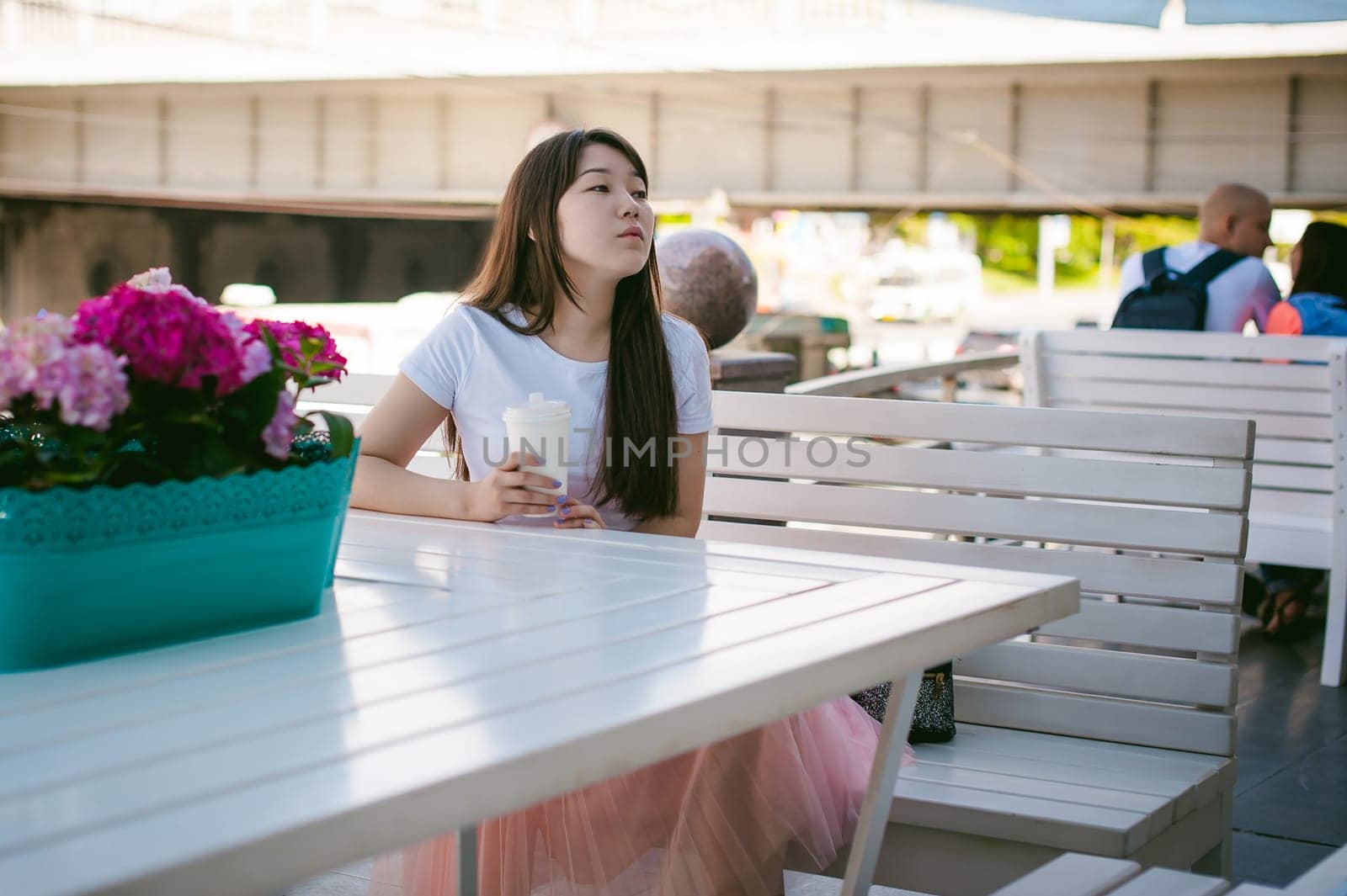 Cute asian young woman in summer cafe outdoors. girl In white T-shirt, with long hair in simple light cozy interior of restaurant Urban style, drinking coffee drink, sitting at table with flowers