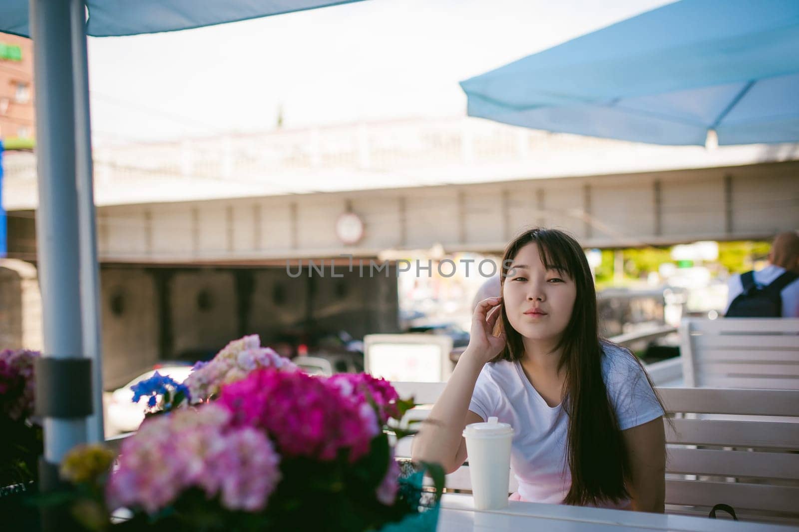 Cute asian young woman in summer cafe outdoors. girl In white T-shirt, with long hair in simple light cozy interior of restaurant Urban style, drinking coffee drink, sitting at table with flowers