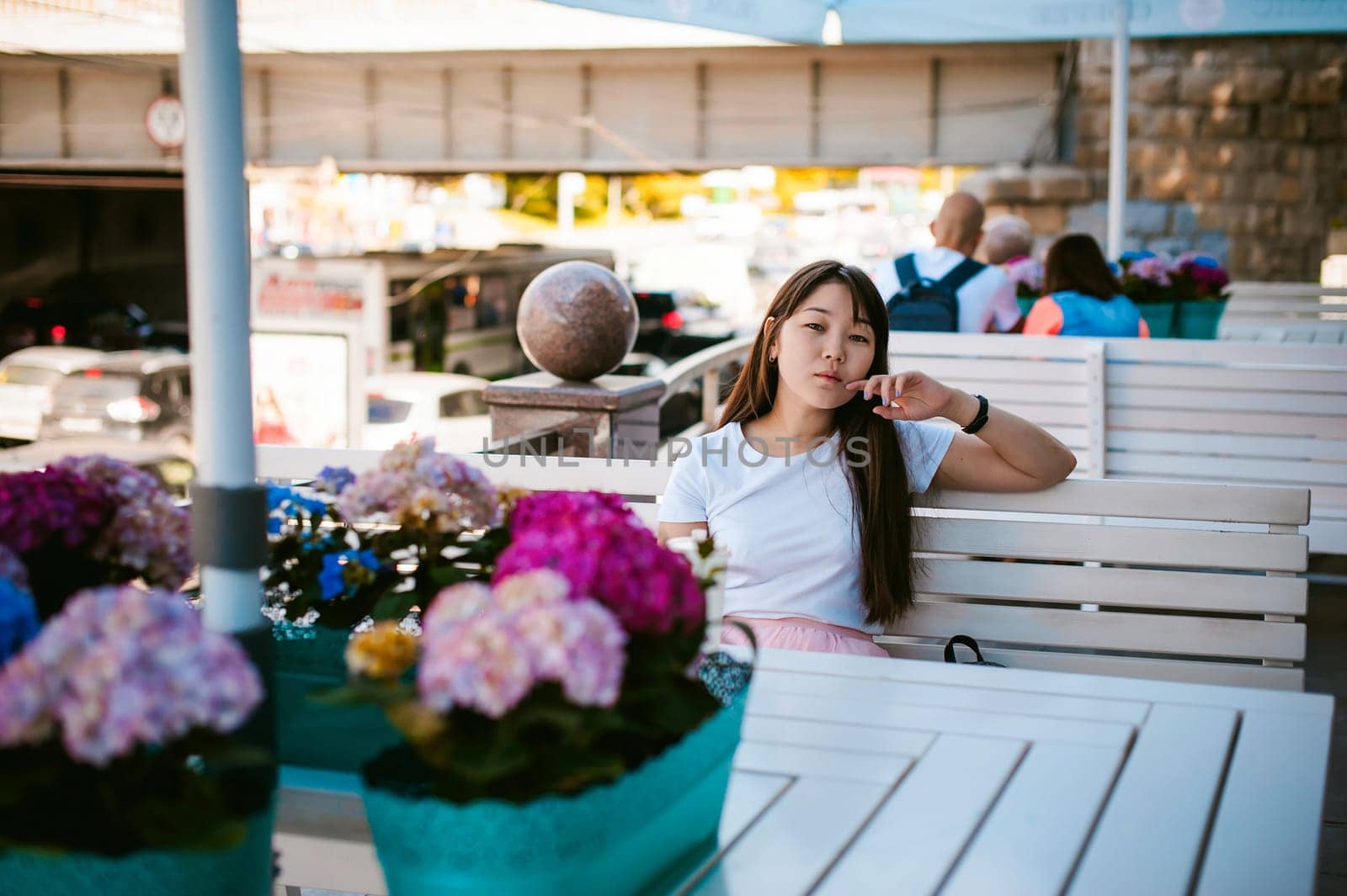 Cute asian young woman in summer cafe outdoors. girl In white T-shirt, with long hair in simple light cozy interior of restaurant Urban style, drinking coffee drink, sitting at table with flowers