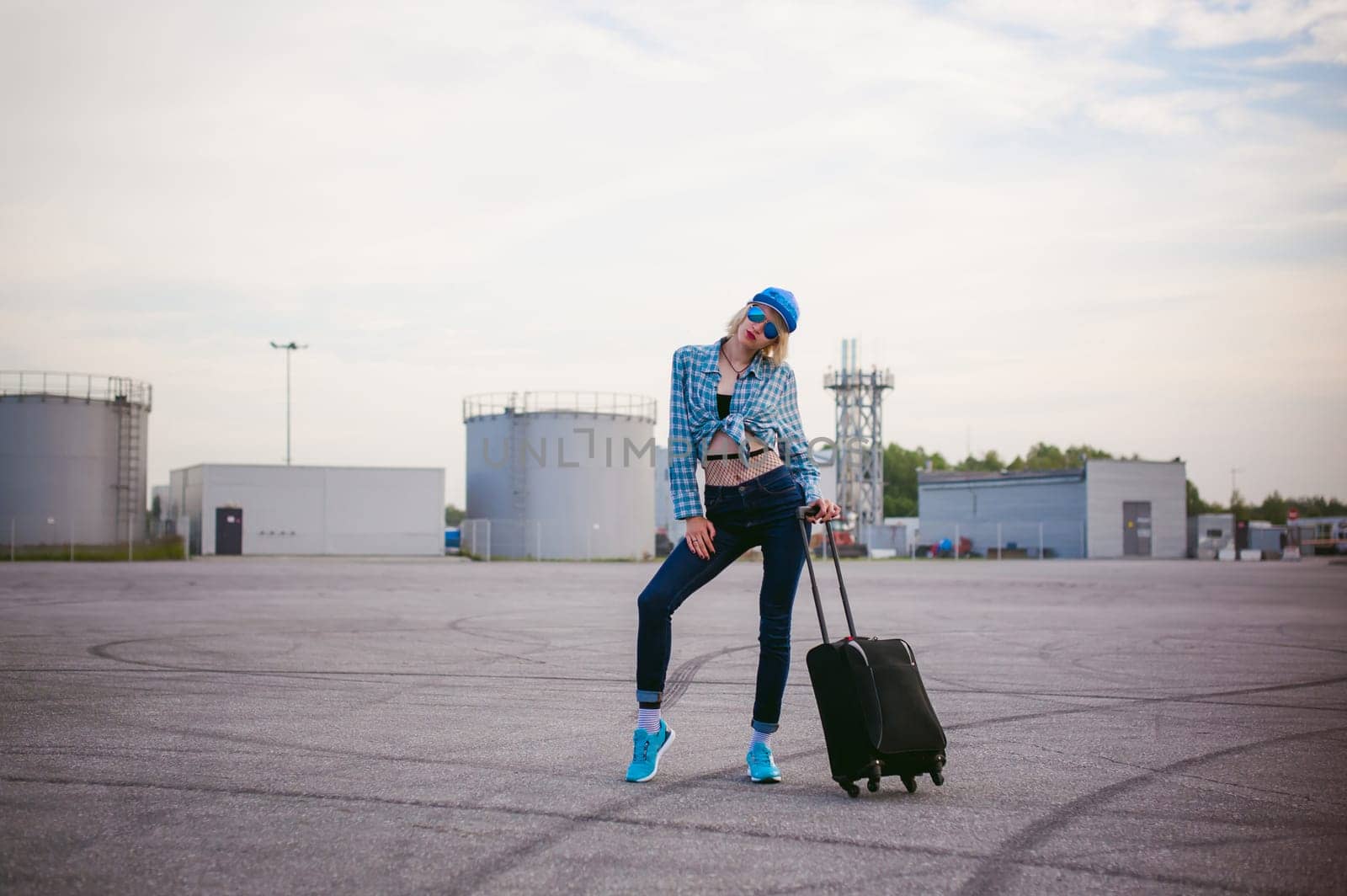young slim woman dressed in blue checked shirt Sunglasses, cap and jeans, Will depart at dawn on summer day on freedom trip with black travel suitcase